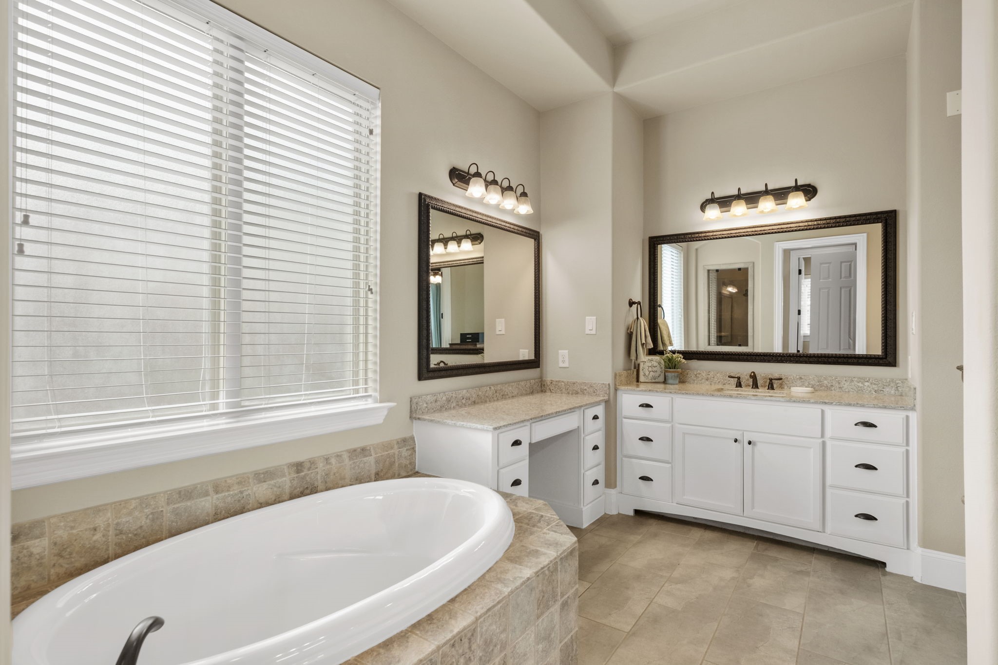 Primary bathroom view showcasing a deep oval soaking tub set in a custom tile surround beneath a large privacy-glass window, alongside a built-in makeup vanity area with granite counter and drawers.
