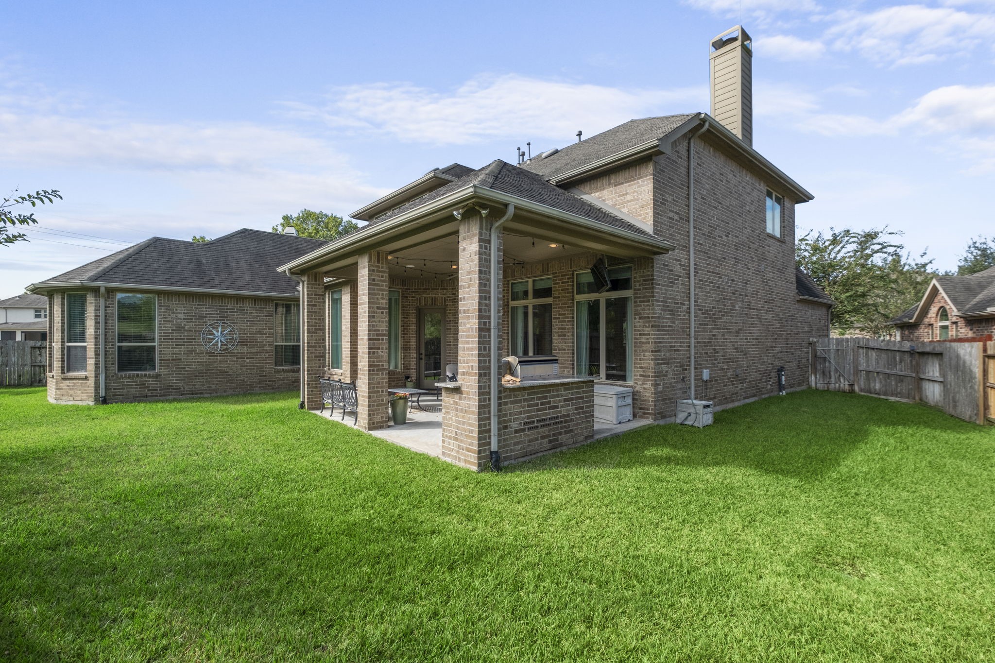Full rear view of the two-story brick home, showcasing the integrated covered patio, outdoor kitchen structure, stone chimney stack, and gabled roofline.