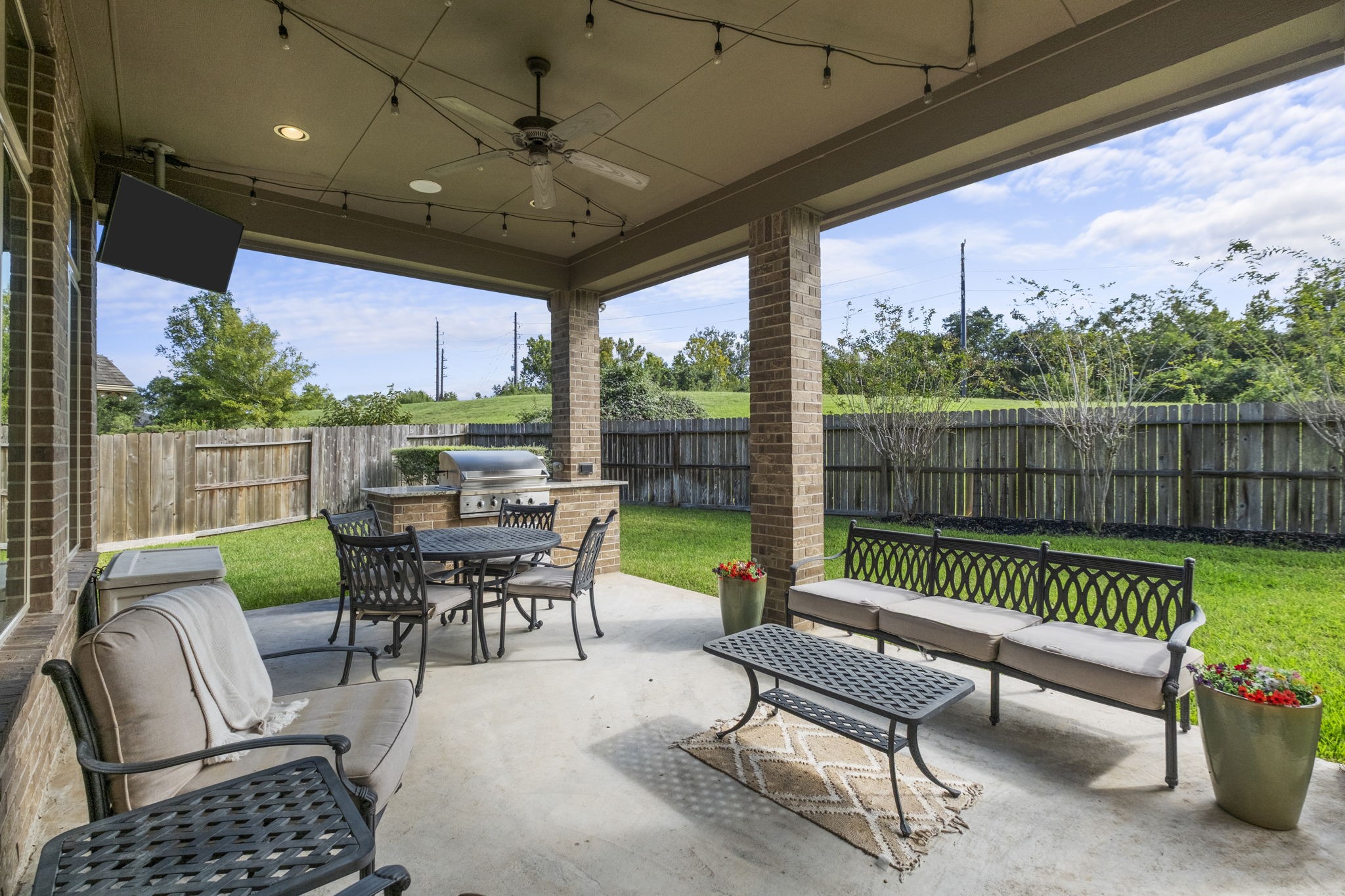Spacious covered back patio featuring a beadboard ceiling with a ceiling fan, recessed lighting, and outdoor string lights. Equipped with a built-in outdoor kitchen with granite countertops, integrated grill, outdoor TV mount, and stone support pillars.