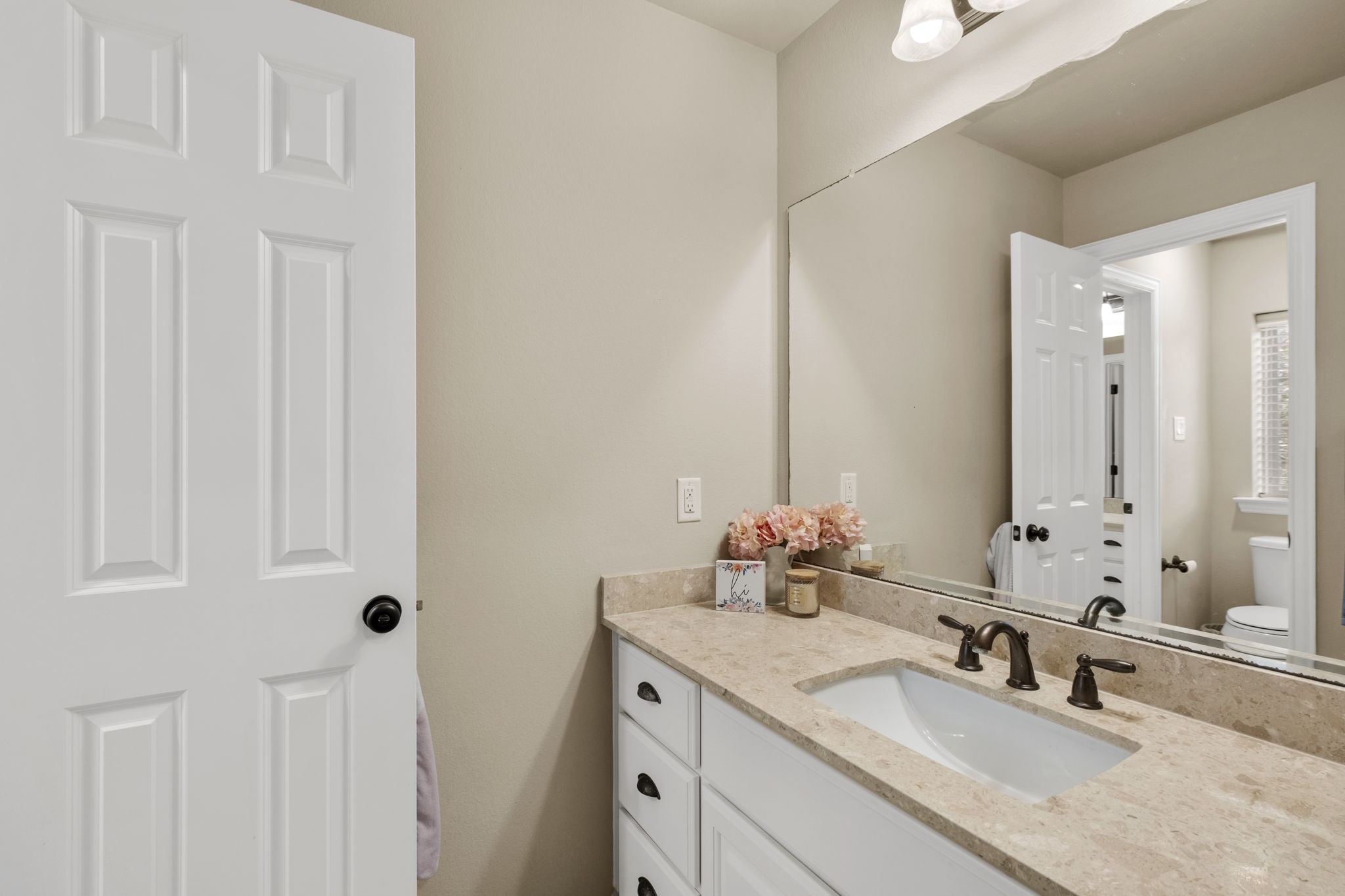 Detail view of the second-story bathroom vanity space showcasing white raised-panel cabinetry, dark drawer pulls, oil-rubbed bronze plumbing fixtures, neutral wall finish, and a wide plate-glass vanity mirror reflecting light from the adjacent hallway and bedroom.