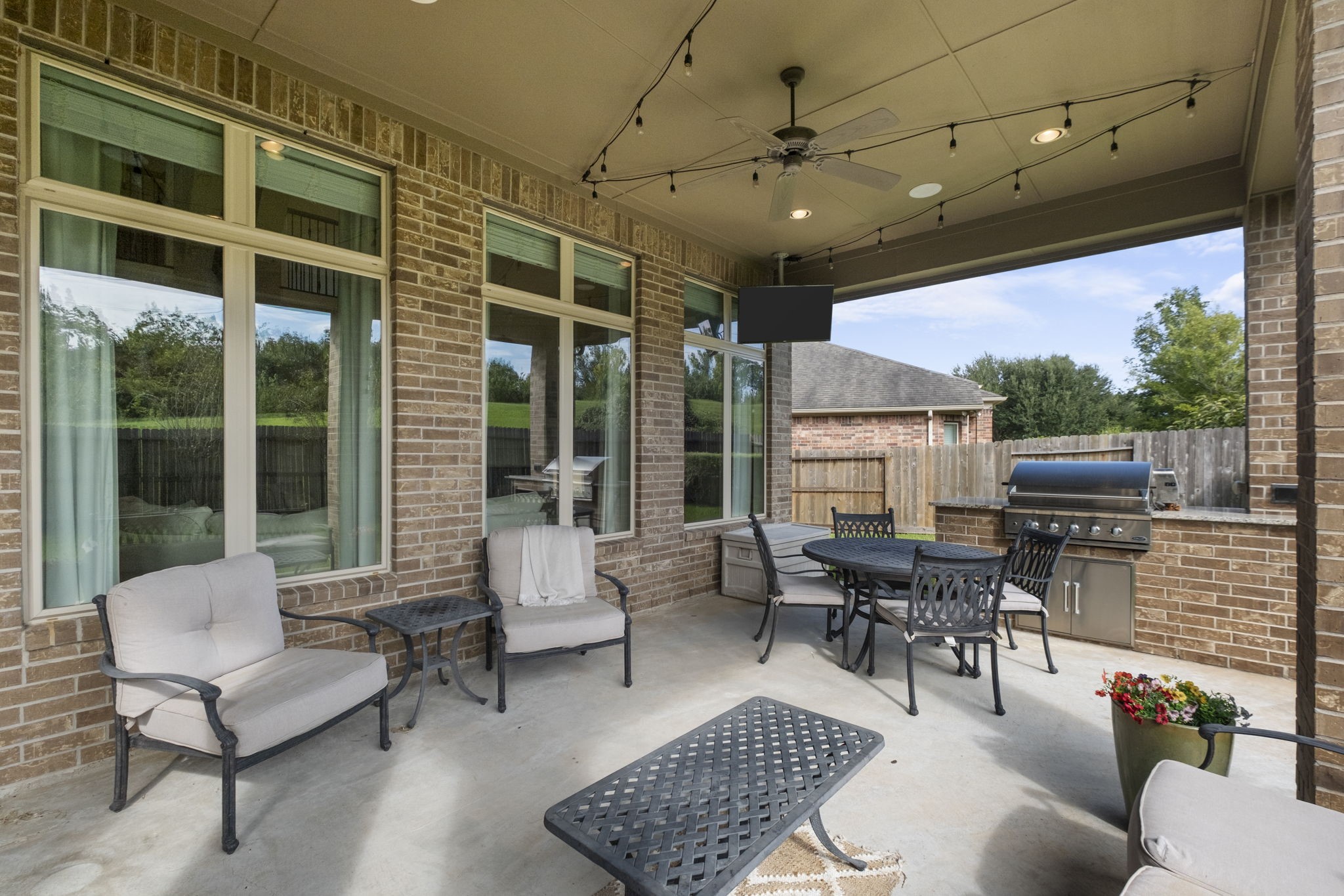Full view of the covered patio area showing floor-to-ceiling multi-panel rear windows and glass doors providing seamless indoor-outdoor sightlines. Features a built-in stainless steel BBQ grill with granite counter space and a brick base.