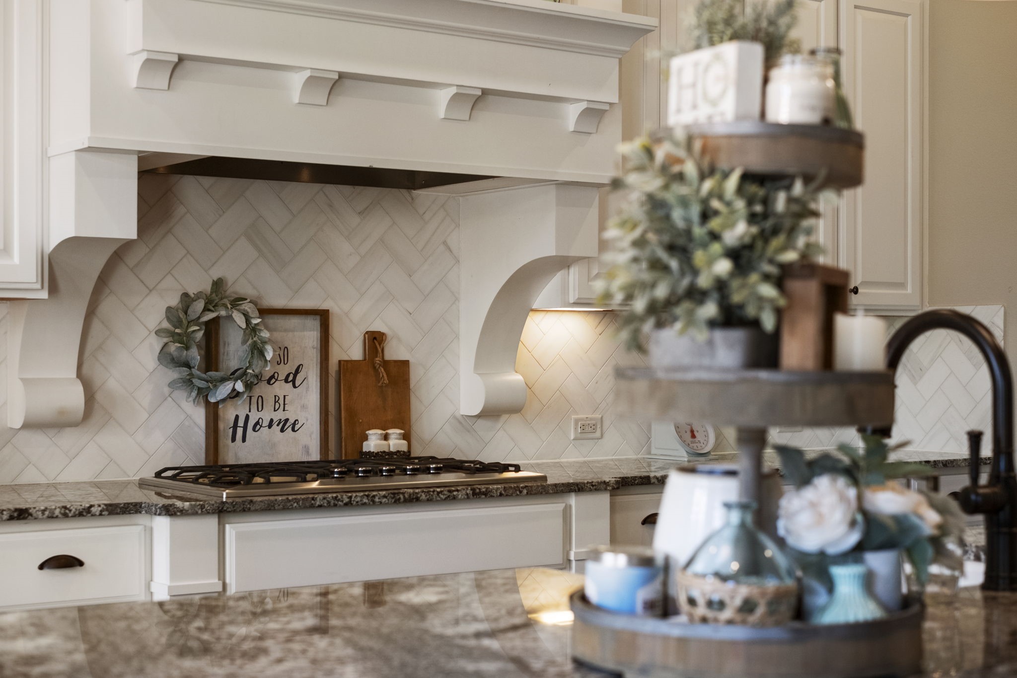 Detailed view of the custom white-painted wood range hood featuring carved corbels, built over a gas cooktop set in granite counter space. Highlights the light-toned subway tile arranged in a classic herringbone backsplash pattern with under-cabinet accent lighting.