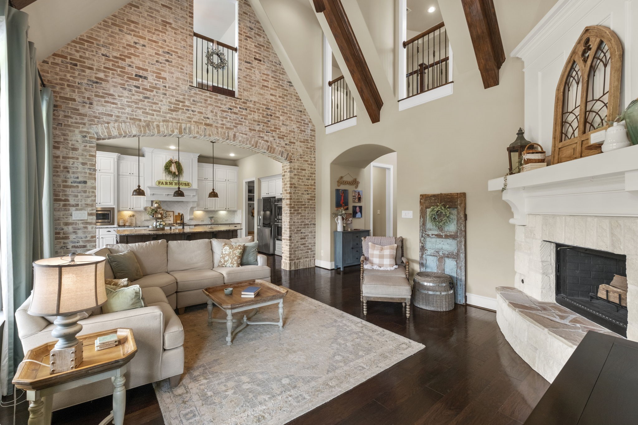 High-angle living room perspective showcasing the full double-height ceiling, upper-level hallway overlooks with wrought-iron railings, exposed wood beams, and a full accent wall crafted in exposed brick with an oversized architectural archway.