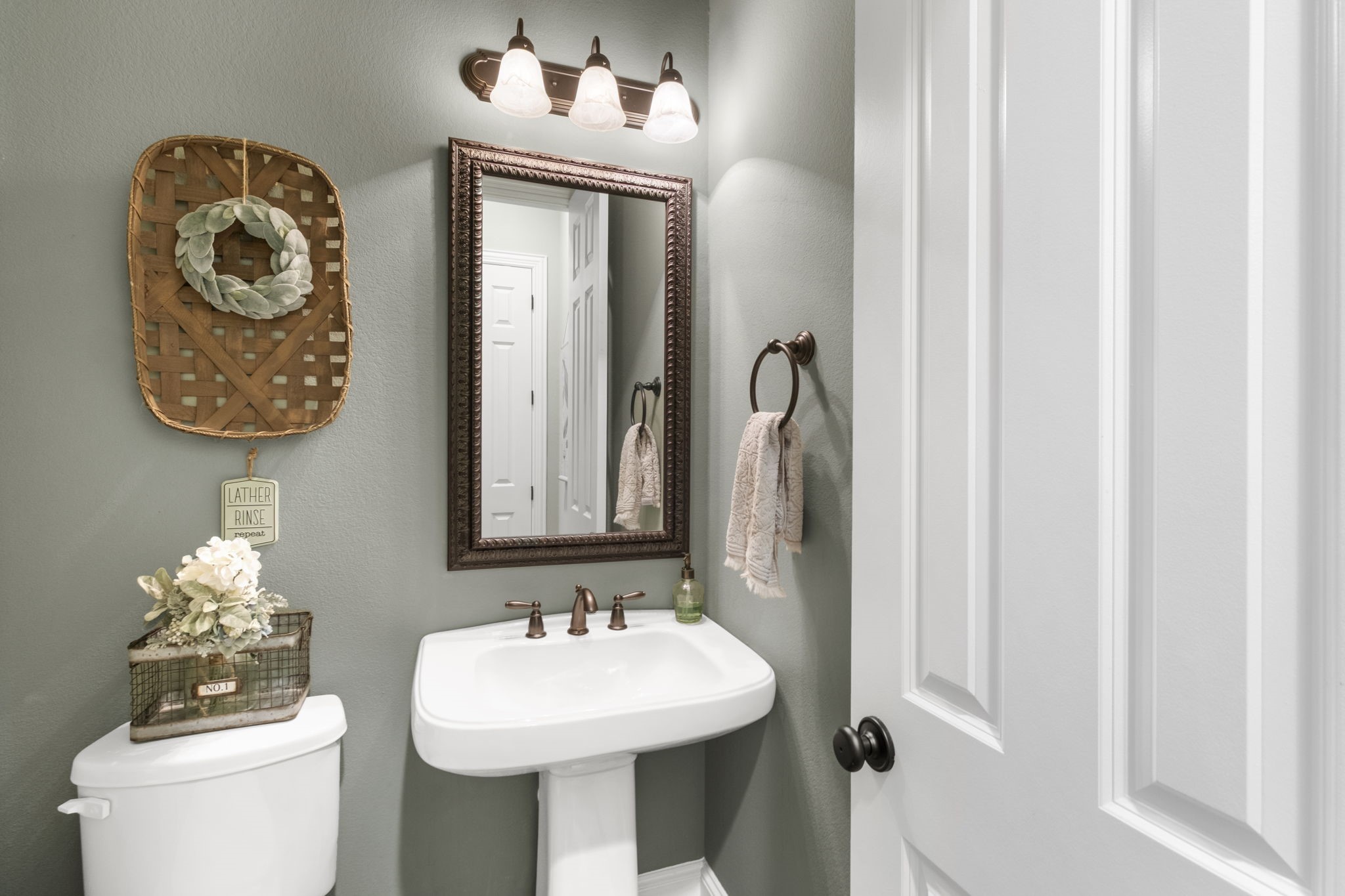 Charming half-bath featuring a classic white porcelain pedestal sink with bronze fixtures, oil-rubbed bronze vanity lighting, decorative framed mirror, neutral sage green walls, and tile flooring.