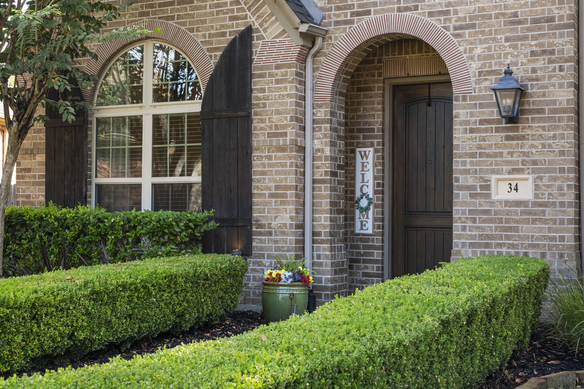 Close-up exterior view highlighting the home's detailed brickwork, double arched entryways with brick header accents, rustic wooden shutters on the multi-pane arched window, a lantern light fixture, and sculpted boxwood hedging.