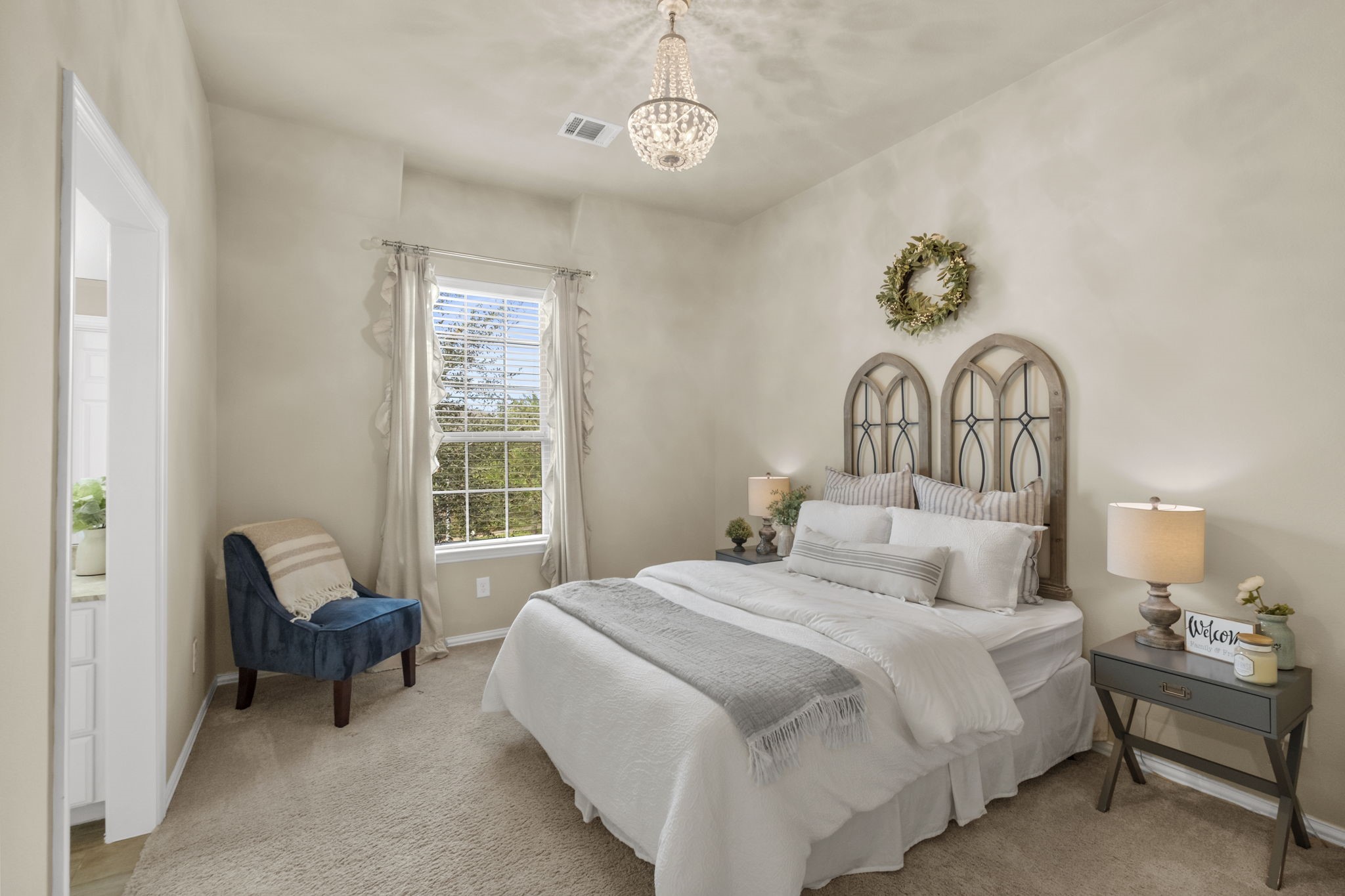 Sunlit guest bedroom featuring plush carpeting, a vaulted ceiling line, an elegant crystal chandelier light fixture, neutral walls, and a large window overlooking the outdoor grounds. Direct access leads to an adjacent bathroom.
