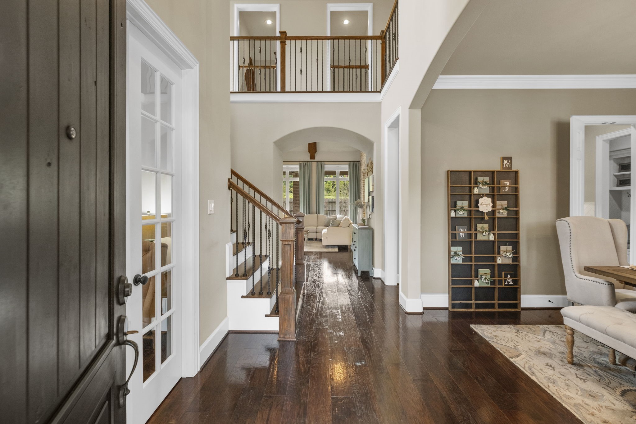 Grand two-story foyer entry featuring warm hardwood flooring, a sweeping staircase with wrought-iron spindles, recessed lighting, and an upper-level overlook balcony. Archway leads toward the light-filled main living space.