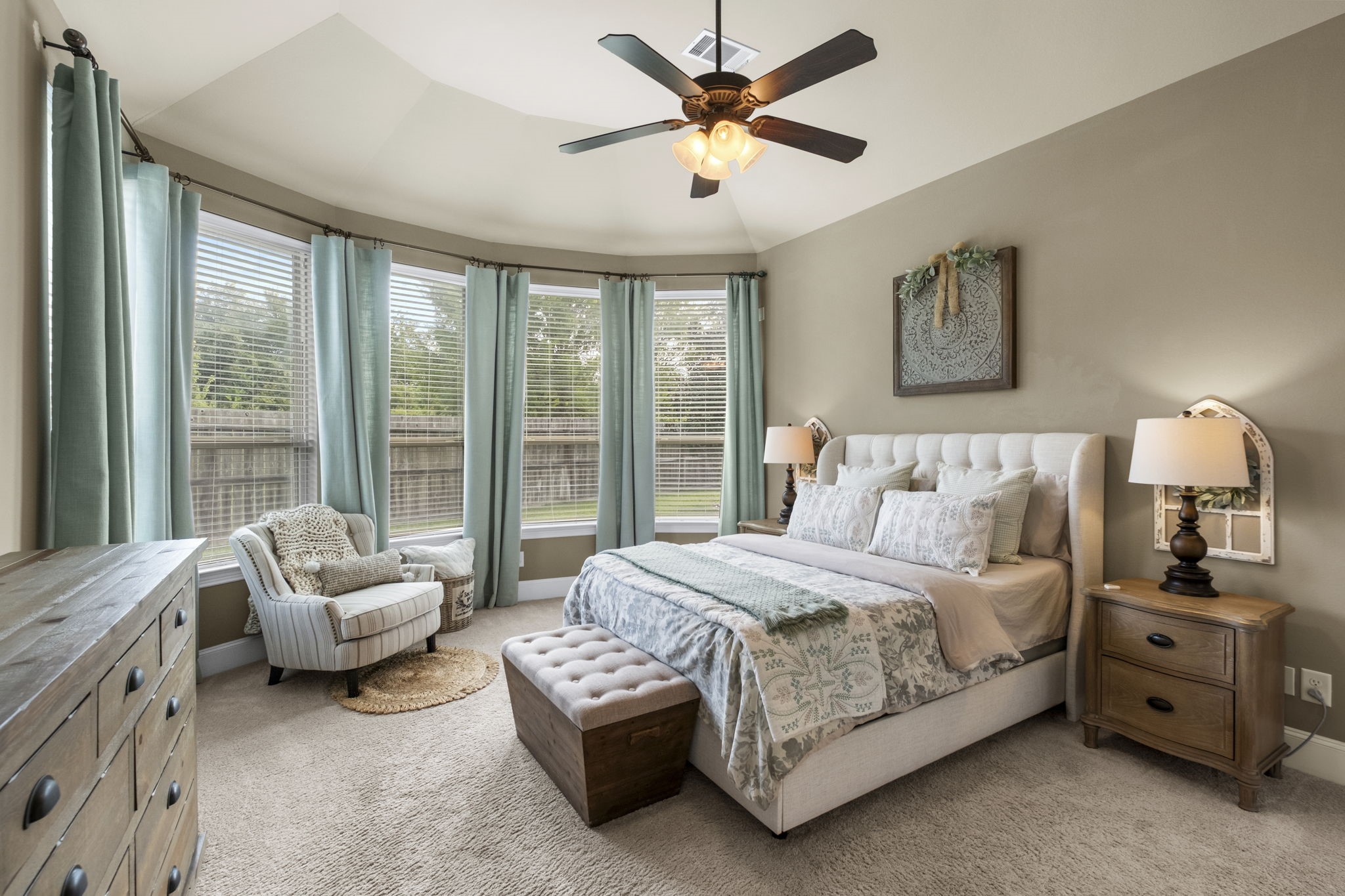 Spacious primary suite featuring plush carpeting, a tray ceiling with a integrated ceiling fan/light fixture, and a curved bay window alcove with custom drapery overlooking the backyard.