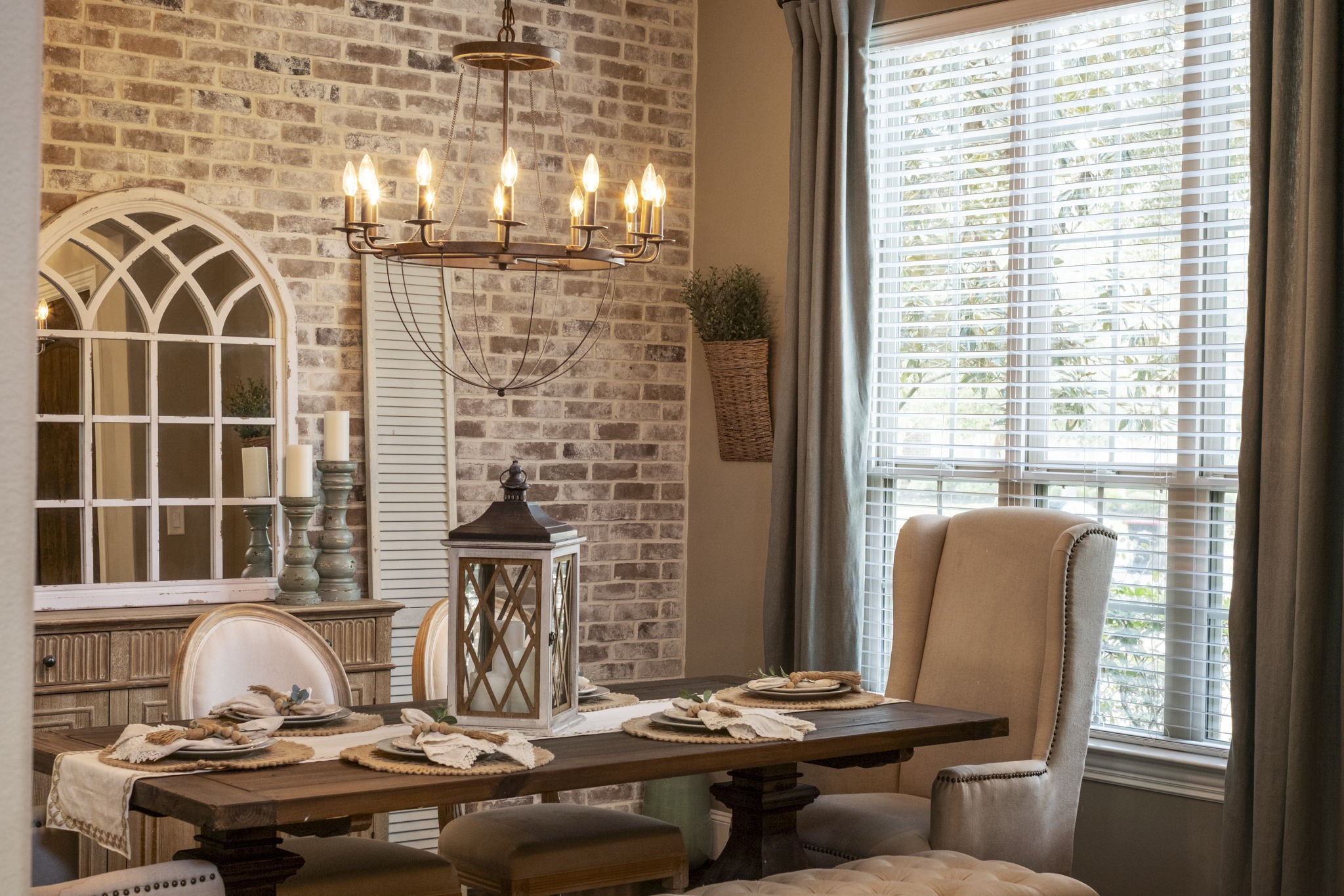 Detailed perspective of the formal dining space highlighting the rustic exposed-brick accent wall, traditional crown molding, dark hardwood floor, and a metal wagon-wheel chandelier. Features a wide double-hung window equipped with trim moulding and blinds.