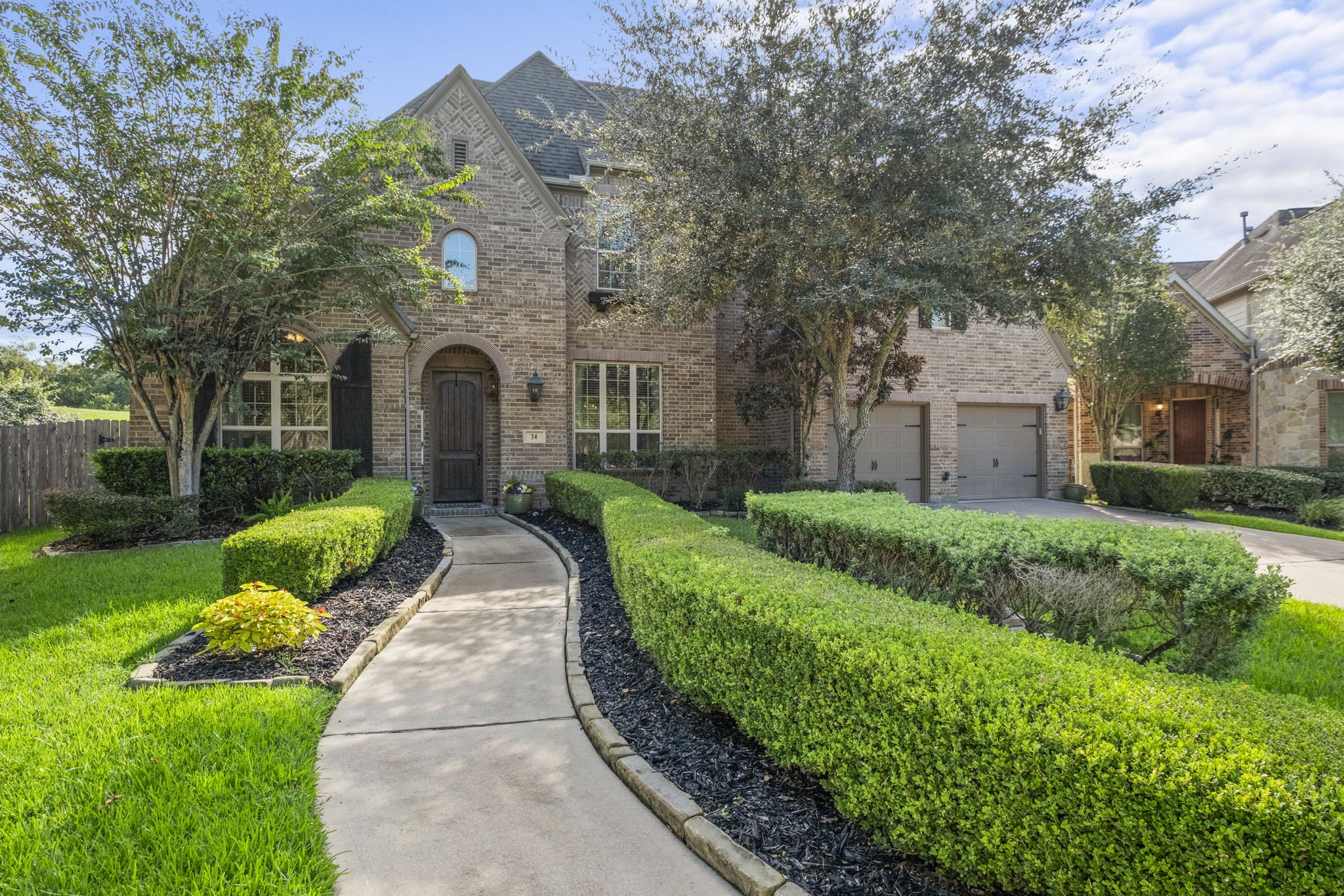Stately two-story brick residence featuring manicured, stone-bordered landscaping, deep mulch beds, and a winding concrete walkway leading to an arched stone entry. Includes double-pane windows with decorative shutters and a tandem multi-car garage.