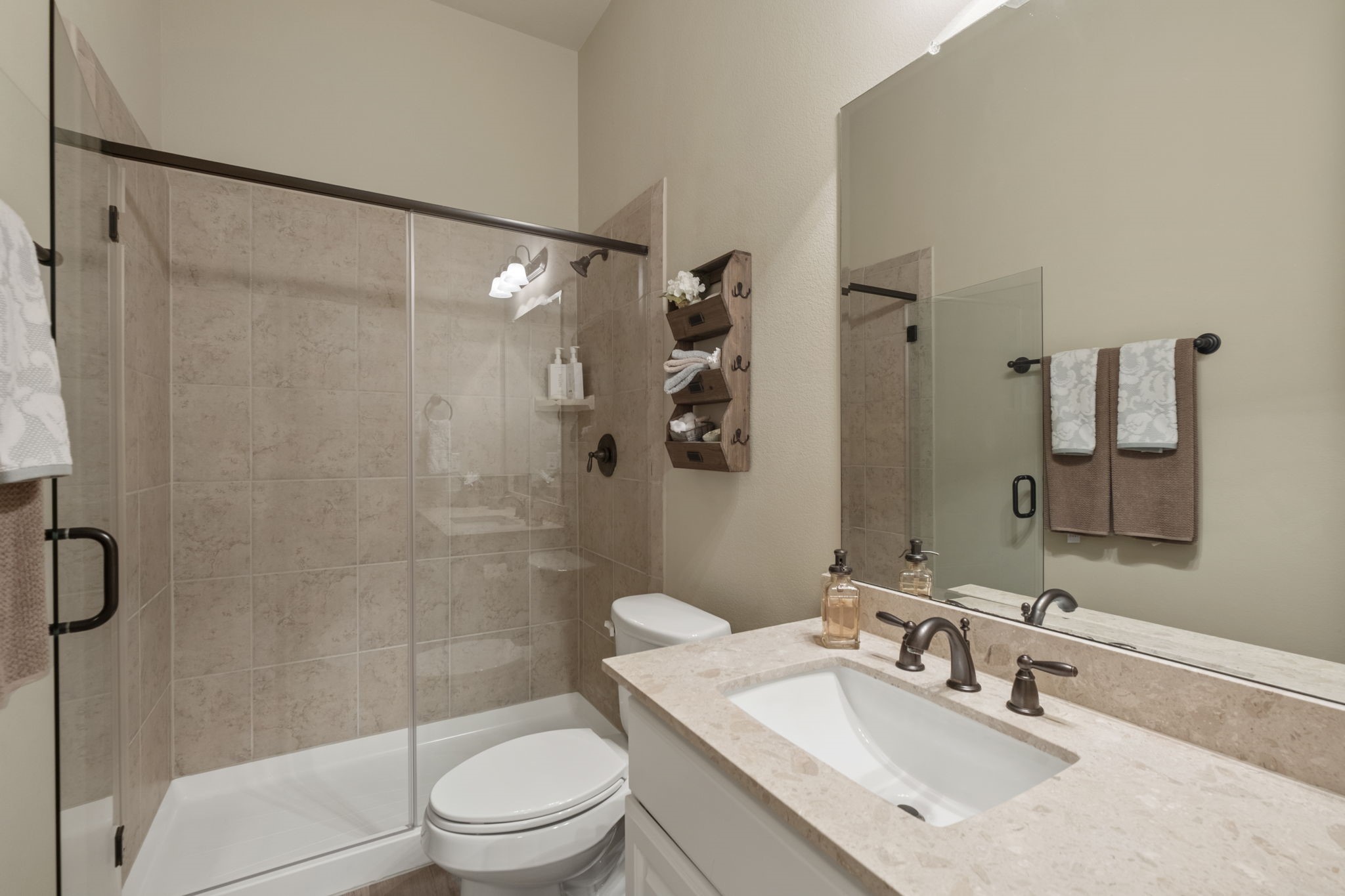 Guest bathroom featuring a neutral tile-surround walk-in shower with a frameless glass sliding door, oil-rubbed bronze fixtures, a toilet, a single-sink vanity with light marble/granite countertops, and a full-width mirror.