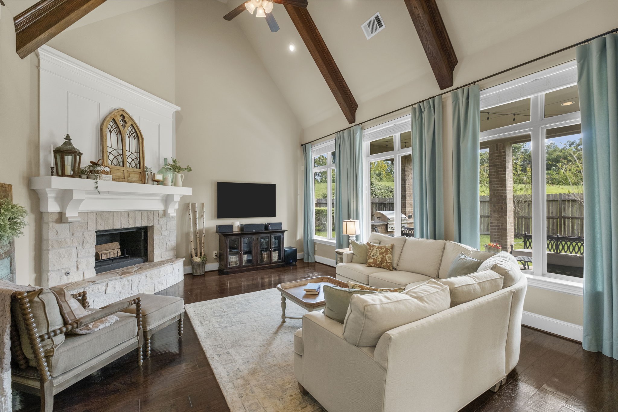 Soaring vaulted ceiling featuring exposed timber beams and large floor-to-ceiling windows overlooking the backyard. The focal point is a floor-to-ceiling floor-mounted stone fireplace with a custom wood mantle and white board-and-batten accent wall above.