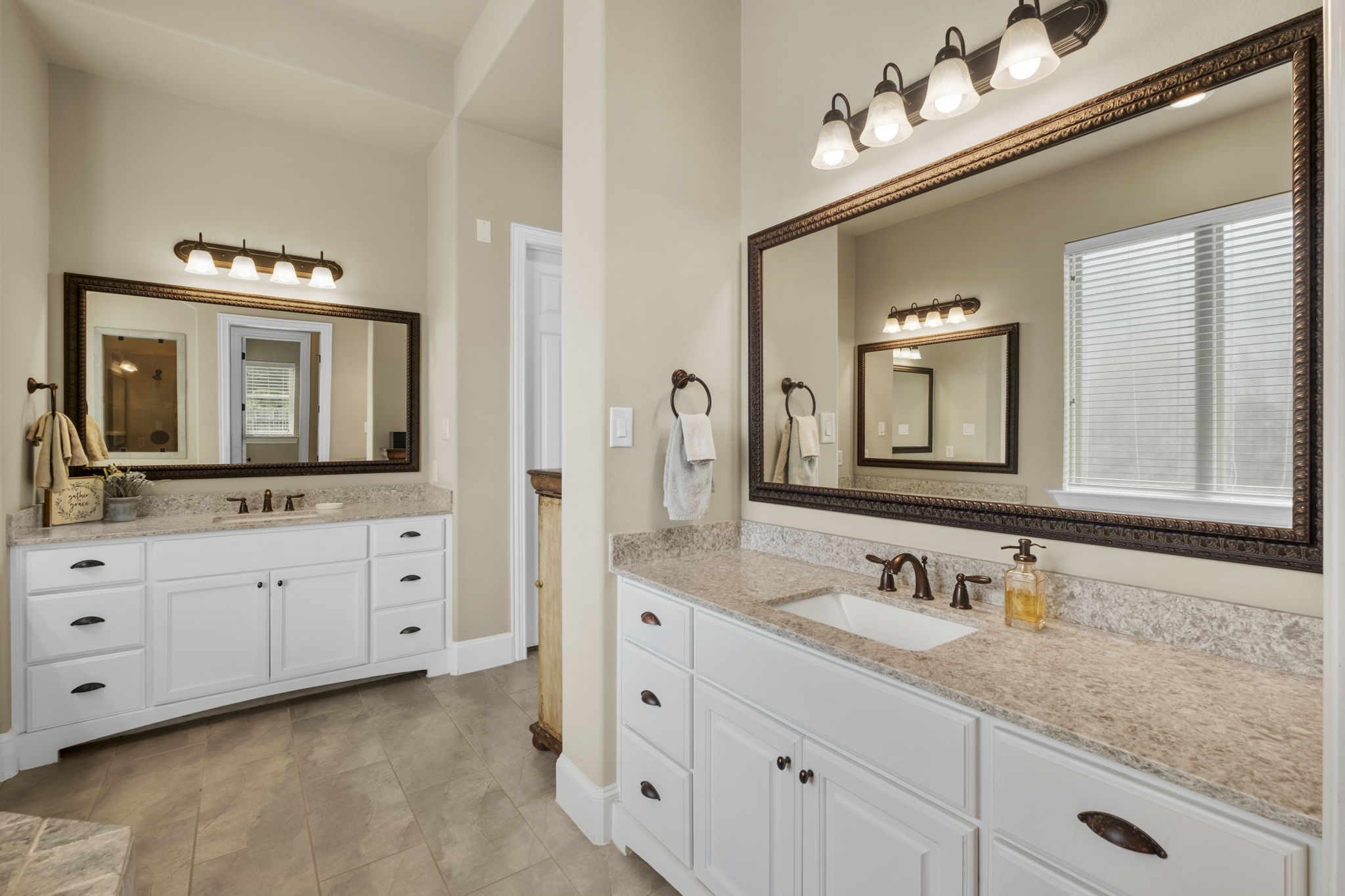 Luxurious primary ensuite featuring dual separated white vanities with granite countertops, oil-rubbed bronze hardware, framed mirrors, updated vanity lights, and neutral tile flooring.