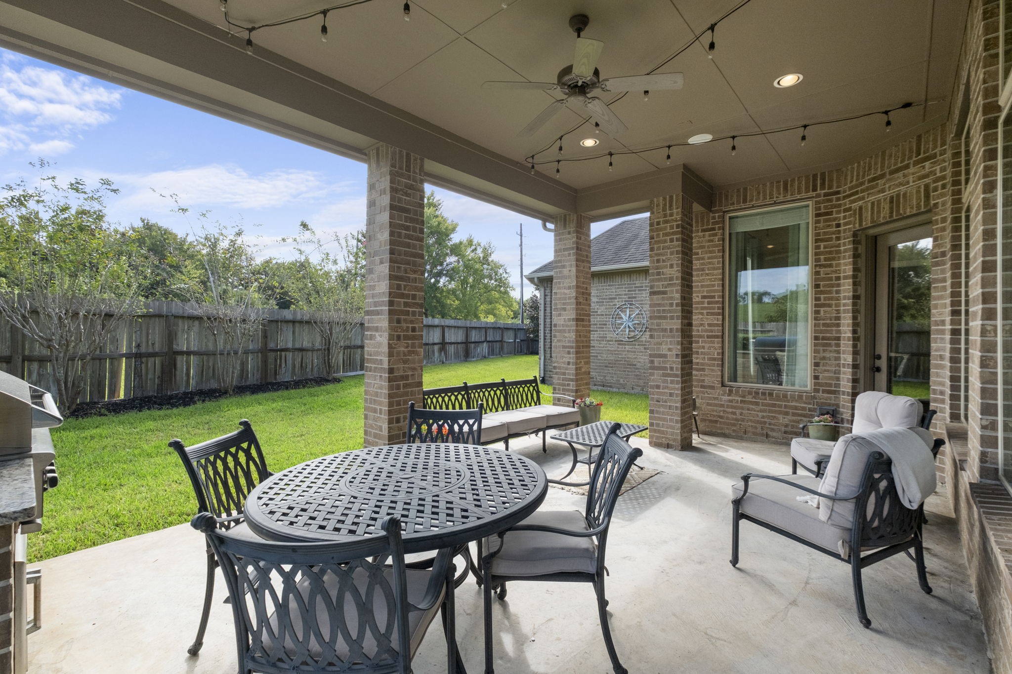 Extended covered patio view highlighting the high-volume ceiling, brick support columns, and concrete deck flooring overlooking the fully fenced green backyard.