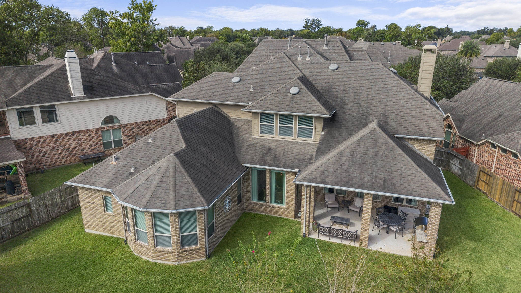 Low-altitude aerial view looking down at the rear of the home, featuring the covered patio, outdoor kitchen area, bay window architectural line, roof dormers, and expansive lawn.