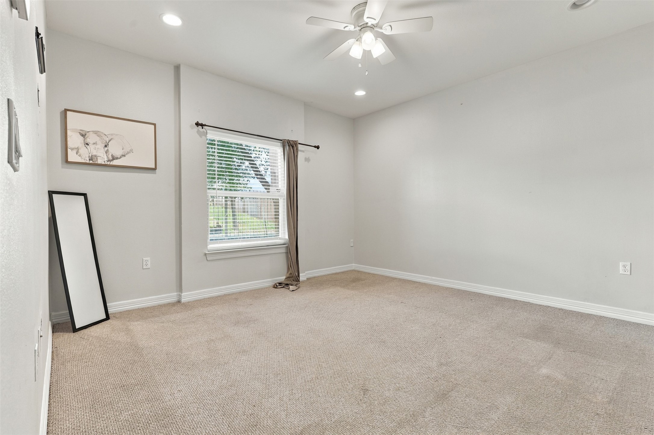 Spacious room with neutral tones, featuring plush carpet, a ceiling fan, and ample natural light from the large window.