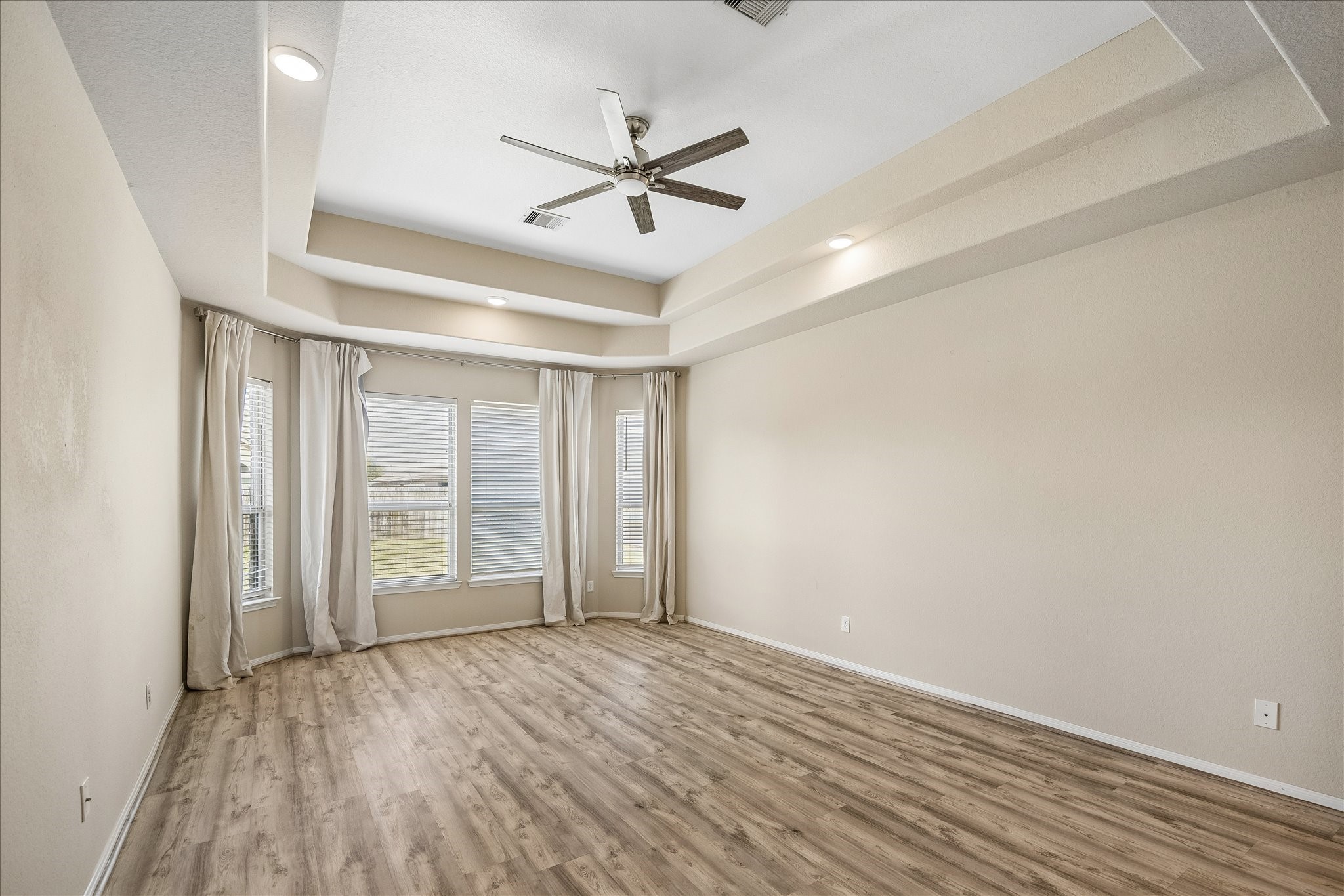 Large primary bedroom with large windows, tray ceiling and wood floors.