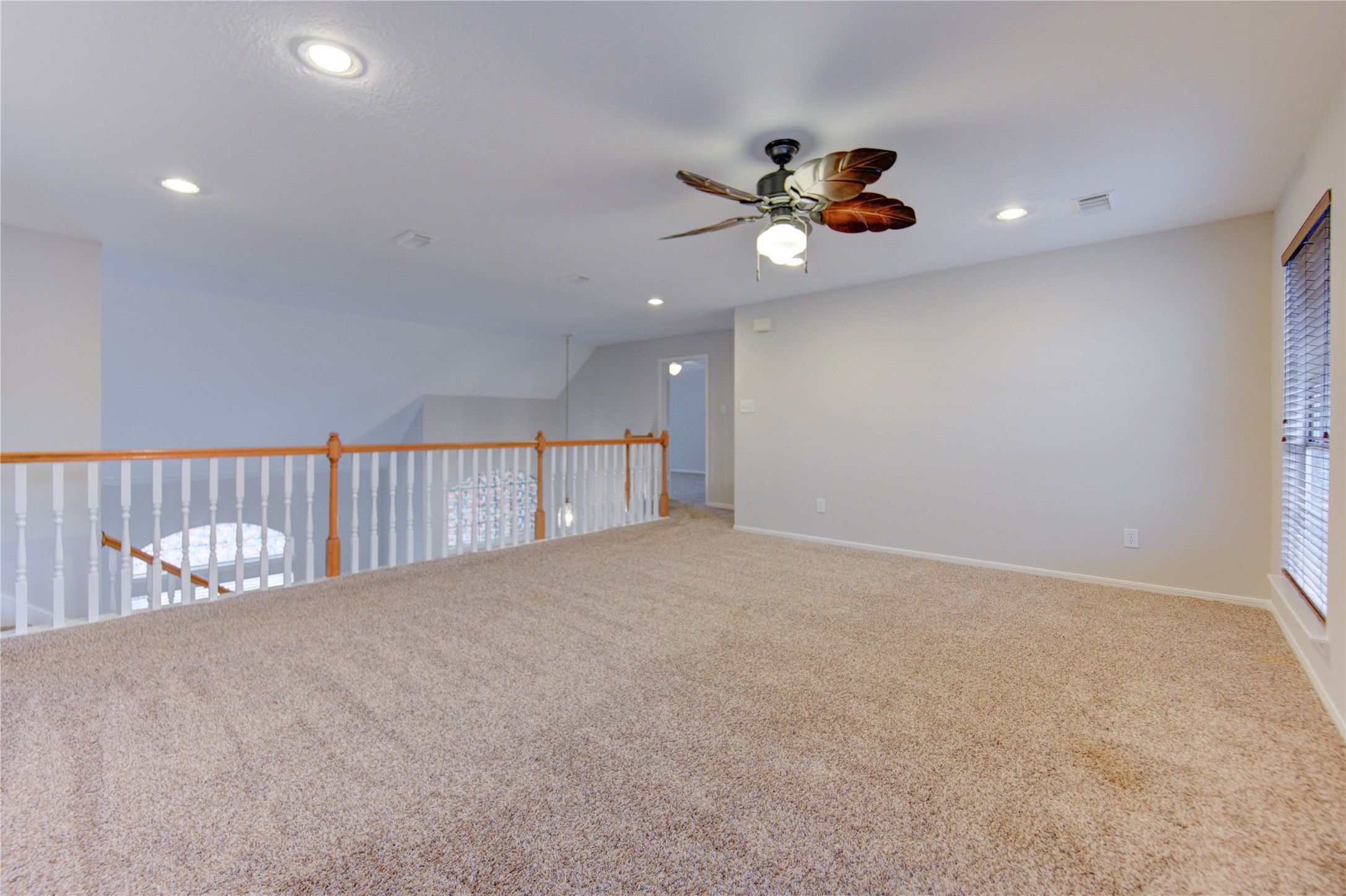 Spacious, carpeted game room with neutral walls, recessed lighting, ceiling fan, two large windows with blinds, and modern wall art, perfect for relaxation or entertaining.