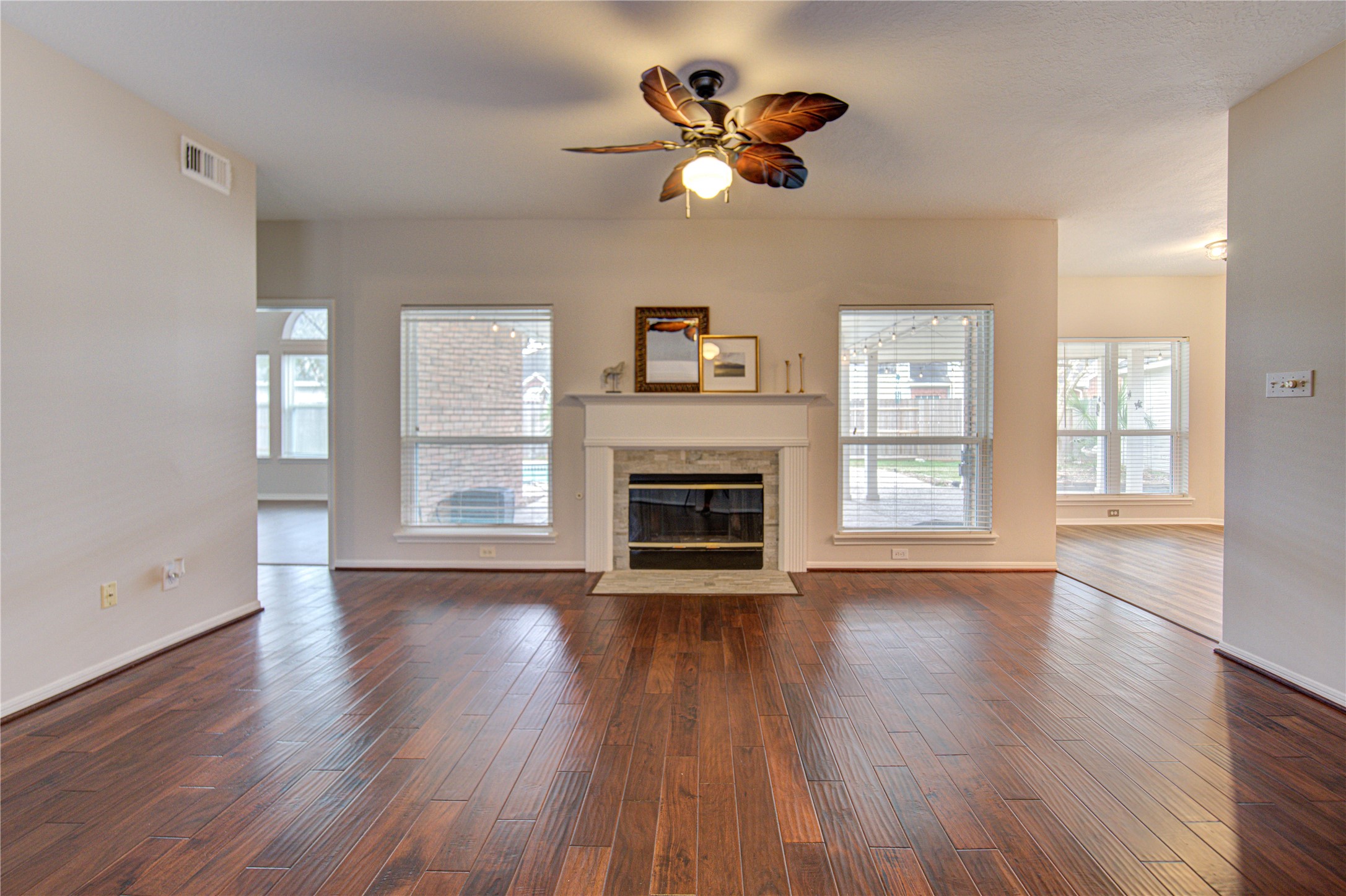 Spacious living room with rich hardwood floors, a cozy fireplace, and large windows offering plenty of natural light. There's a view of a pool outside, and the space is accented with wall decor and a ceiling fan for comfort.
