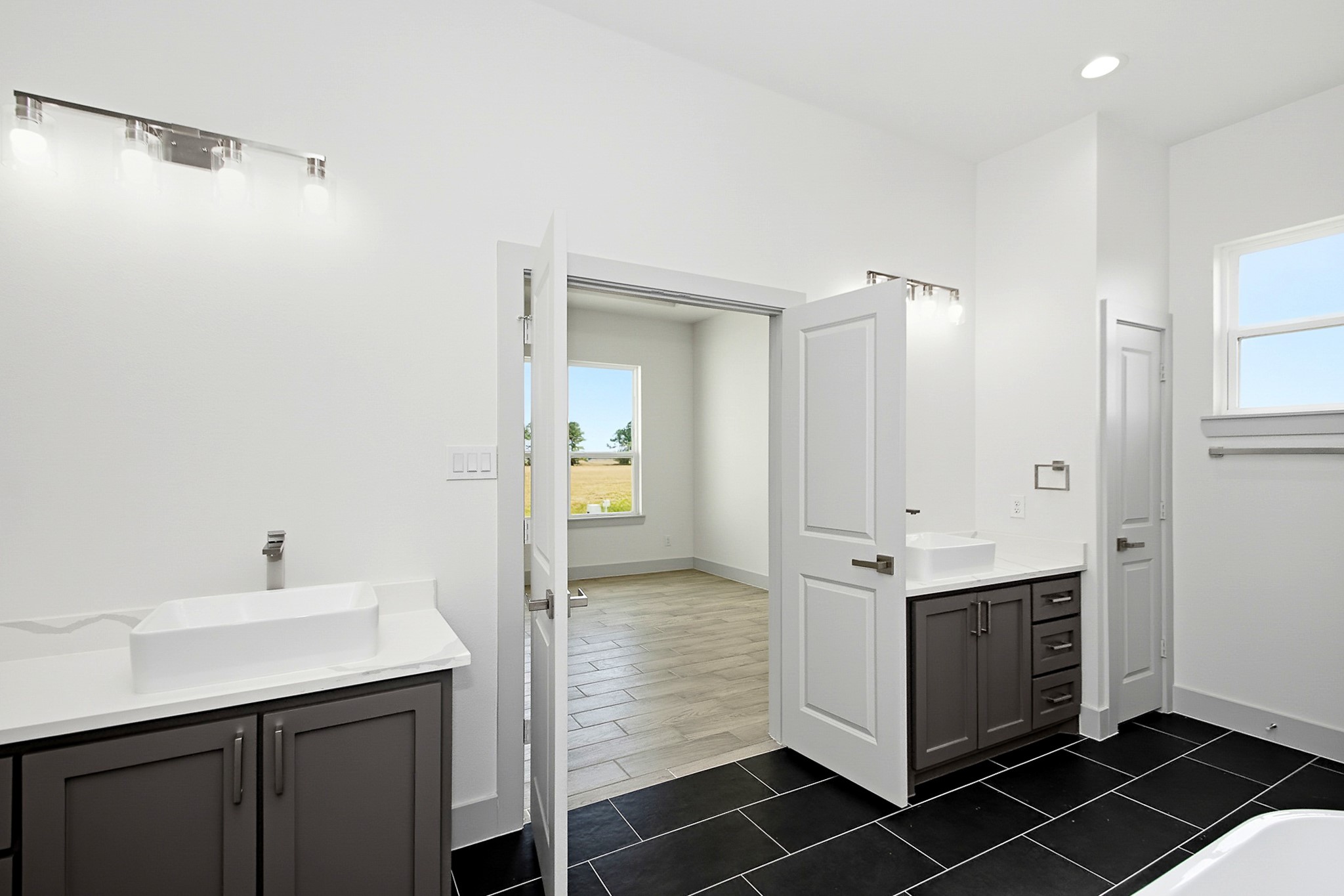 Spacious 16x12 primary bathroom featuring dual vanities, sleek black tile flooring, and ample natural light from large windows, offering a modern and functional design.