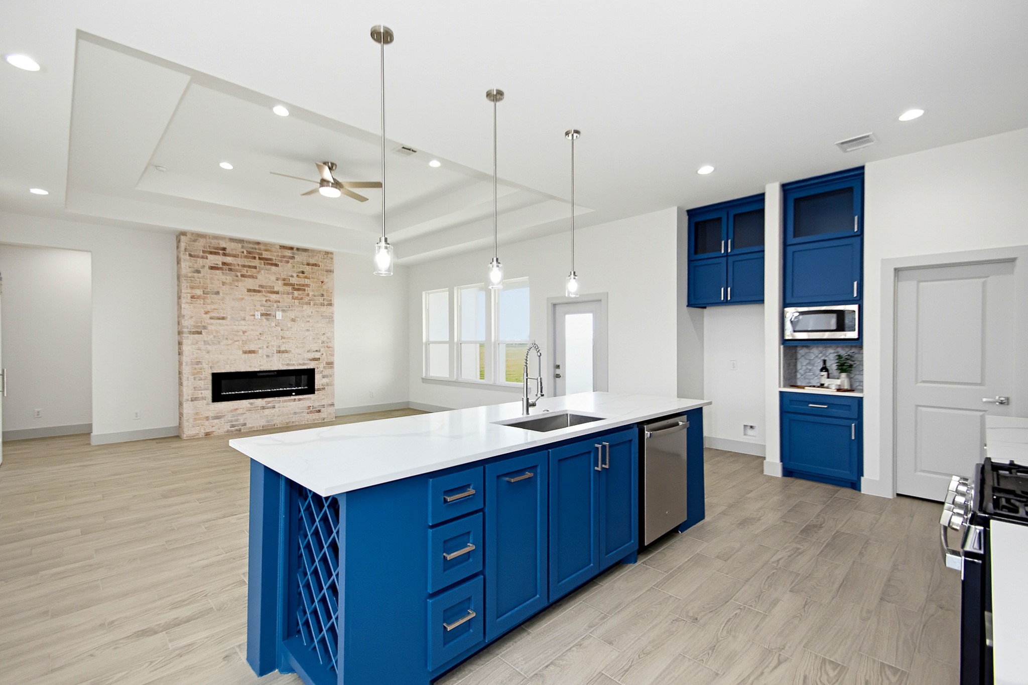Modern 19x14 kitchen with striking blue cabinetry, a spacious island with a built-in wine rack, and elegant pendant lighting. The open-concept living area features a cozy brick fireplace and sleek flooring, enhancing the contemporary ambiance.