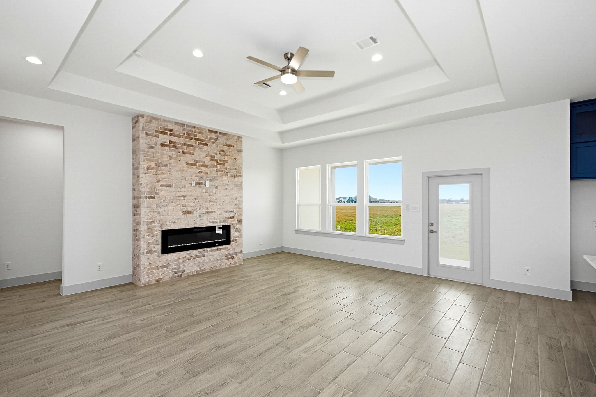 Spacious 19x16 living area with elegant tray ceiling, modern recessed lighting, and a stylish brick fireplace. Large windows and a glass door offer abundant natural light and views of the surrounding landscape.