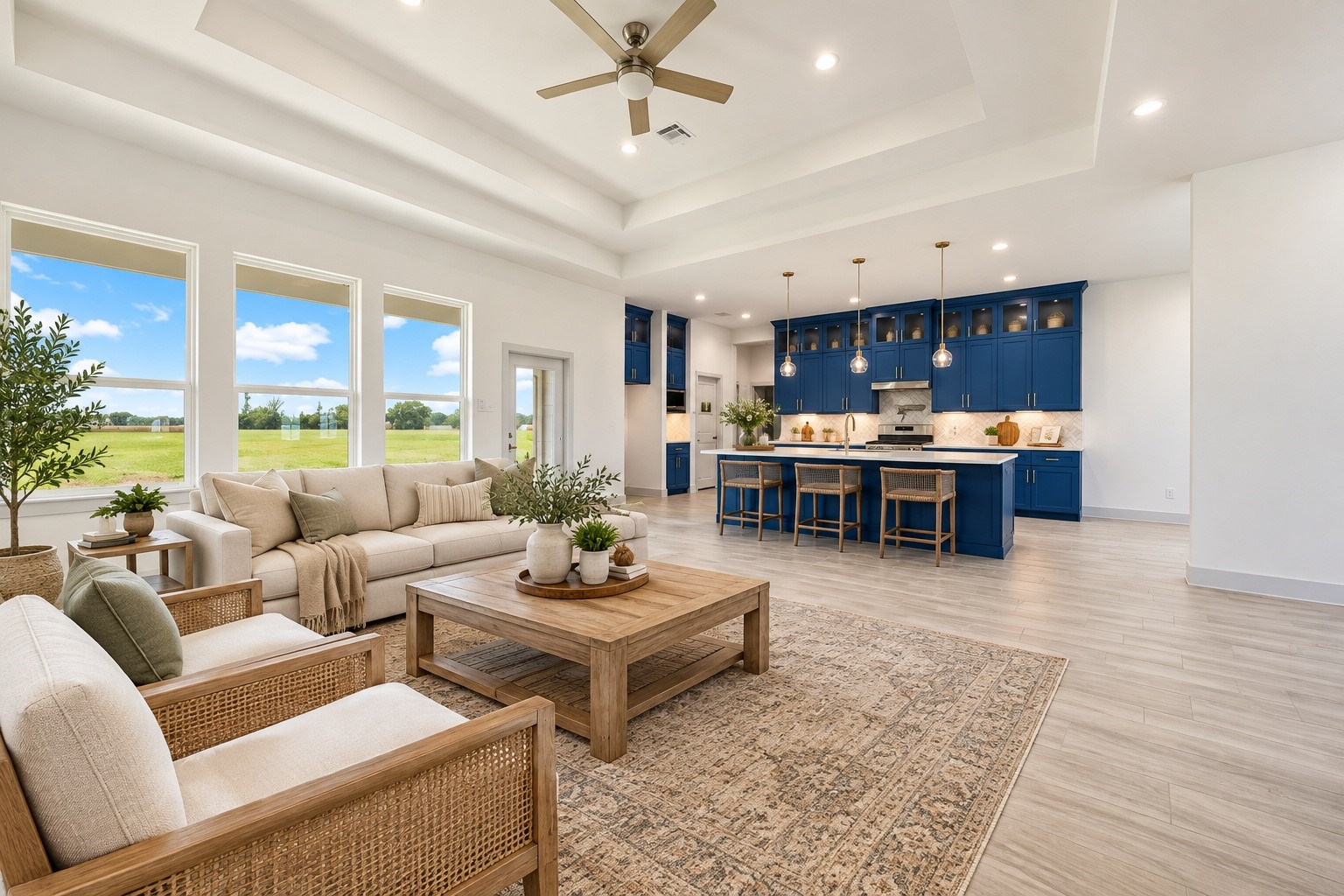 Spacious staged 19x16 open-concept living area with elegant tray ceiling, abundant natural light, and a modern kitchen featuring striking blue cabinetry and a large island.