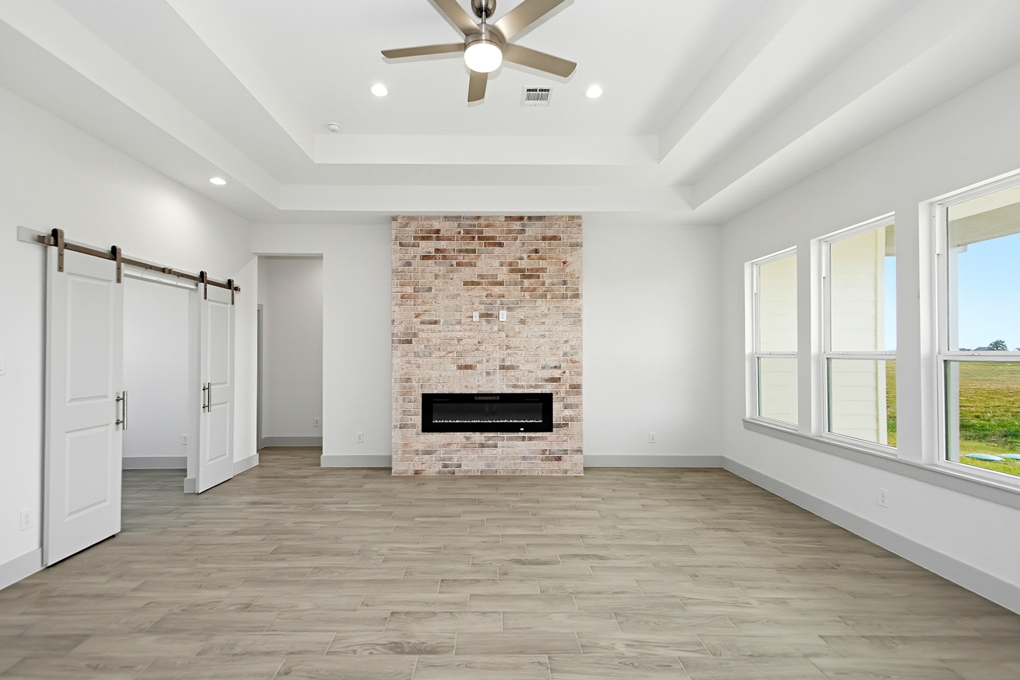 Spacious living area featuring a modern brick fireplace, tray ceiling with recessed lighting, and large windows that fill the room with natural light. Sliding barn doors add a stylish touch to this office space or extra bedroom if needed.