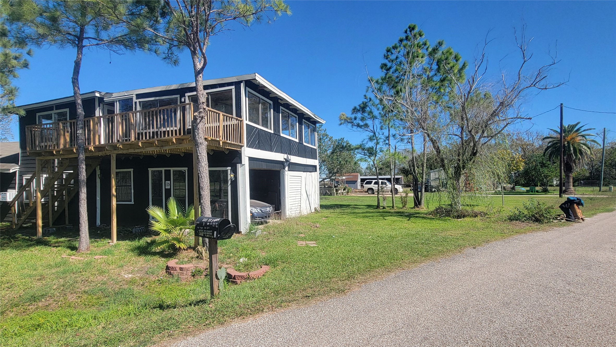 This photo shows a two-story home with a wooden deck and enclosed porch on the upper level. It's surrounded by a grassy yard with trees, positioned on a quiet street. The exterior is a mix of dark and light colors, offering a rustic charm.