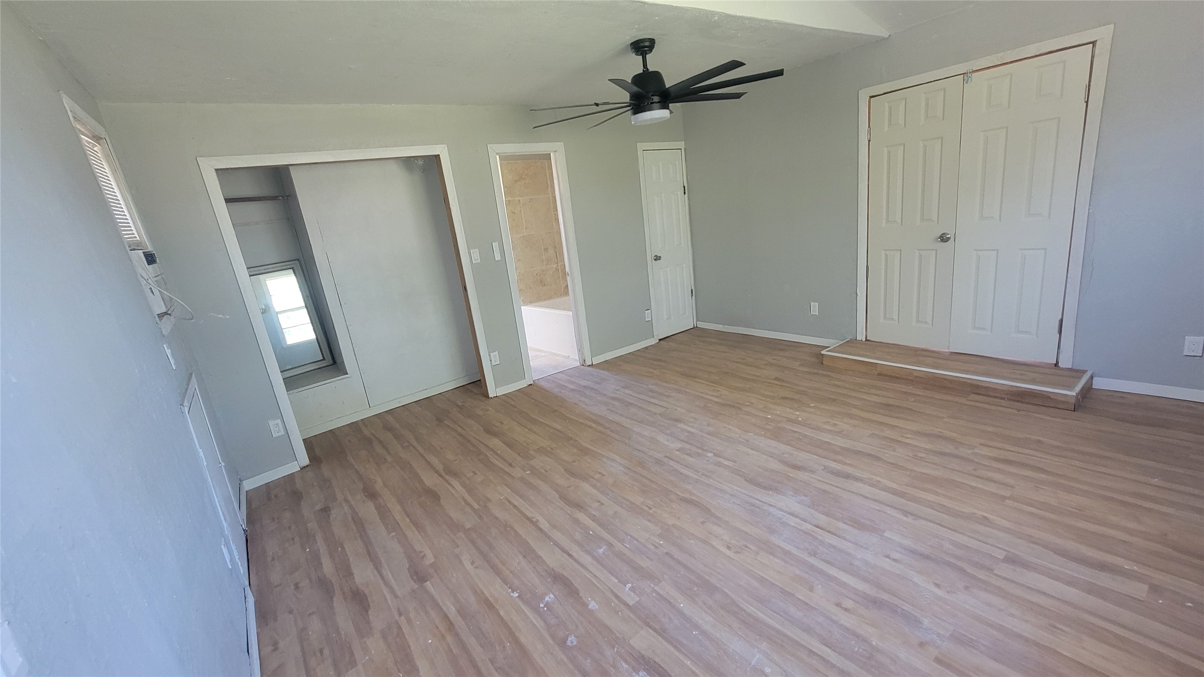 This room features light wood flooring, neutral gray walls, and a modern ceiling fan. It includes a small closet, double doors, and an adjacent bathroom. Ample natural light comes from a side window.