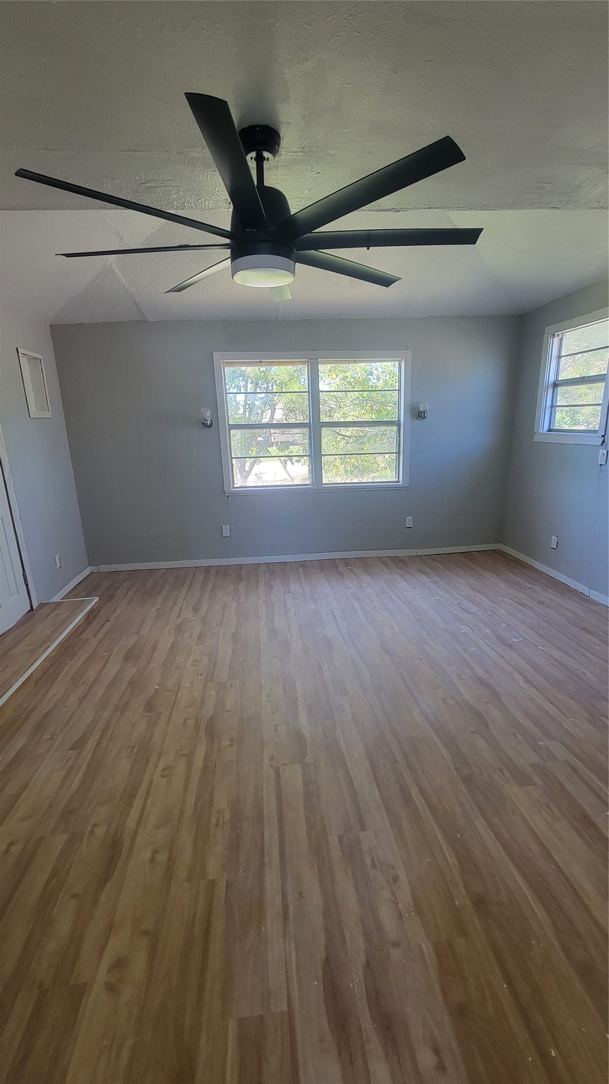 This room features light wood flooring, neutral gray walls, large windows for natural light, and a modern ceiling fan, creating a bright and airy space.