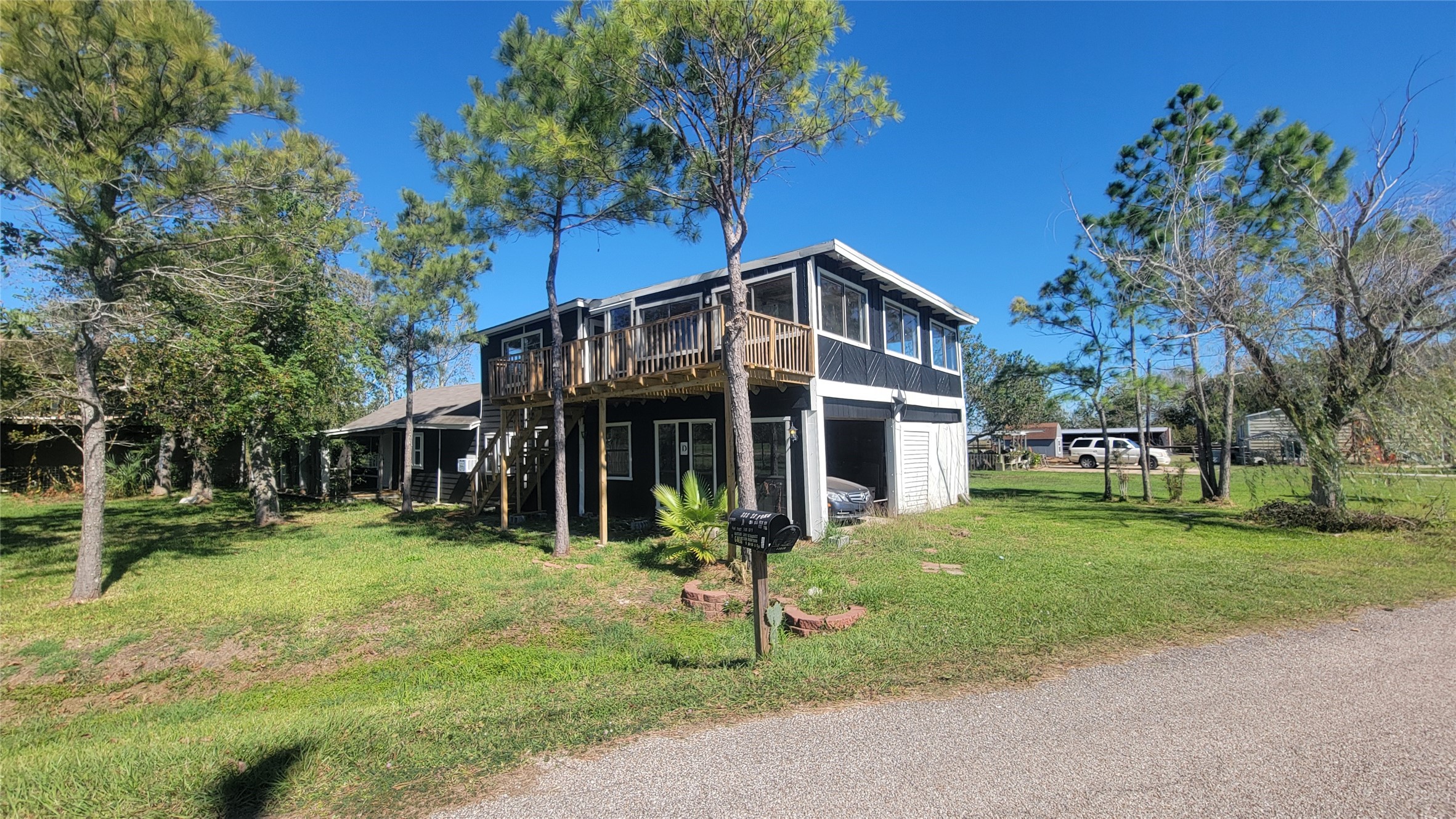 This photo shows a two-story home with a wooden balcony on the upper level, surrounded by trees and grassy areas. The house features a garage and a driveway, set in a quiet, sunny neighborhood.