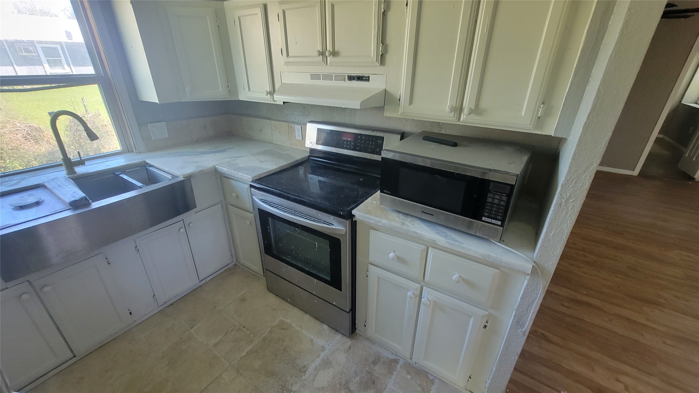 This kitchen features white cabinetry, a stainless steel farmhouse sink, an electric stove, and a microwave. It has a window providing natural light and a view of the backyard. The flooring is tiled, and the adjacent room has light wood flooring.