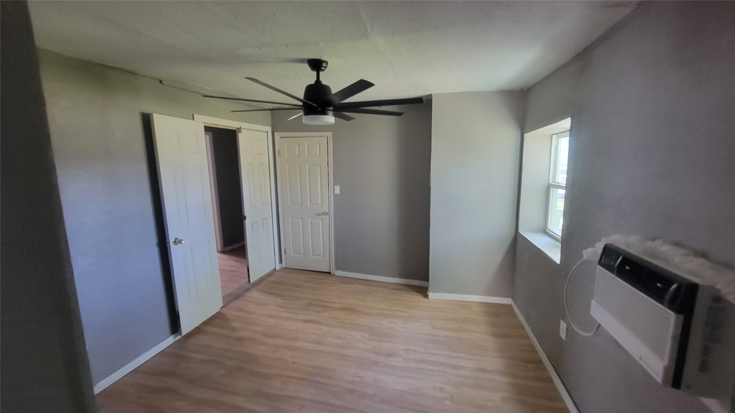 This room features light wood flooring, neutral gray walls, and a ceiling fan. It has a window with a wall-mounted air conditioning unit, providing natural light and ventilation. Two white interior doors lead to adjacent areas.