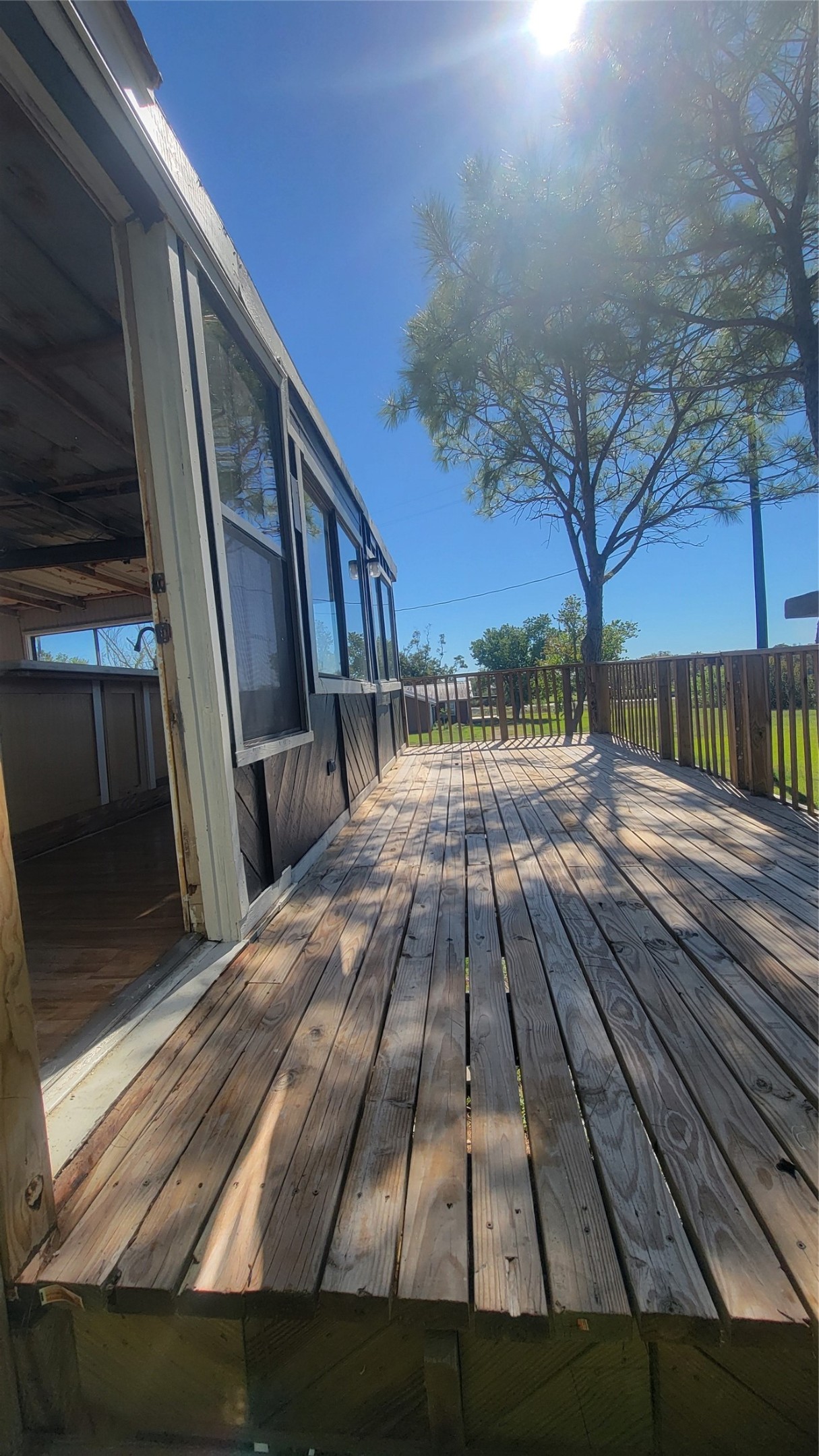 This photo shows a wooden deck attached to a house, partially covered by a porch with large windows. The deck is spacious and surrounded by a railing, with a tree nearby providing some shade. The area looks sunny and inviting.