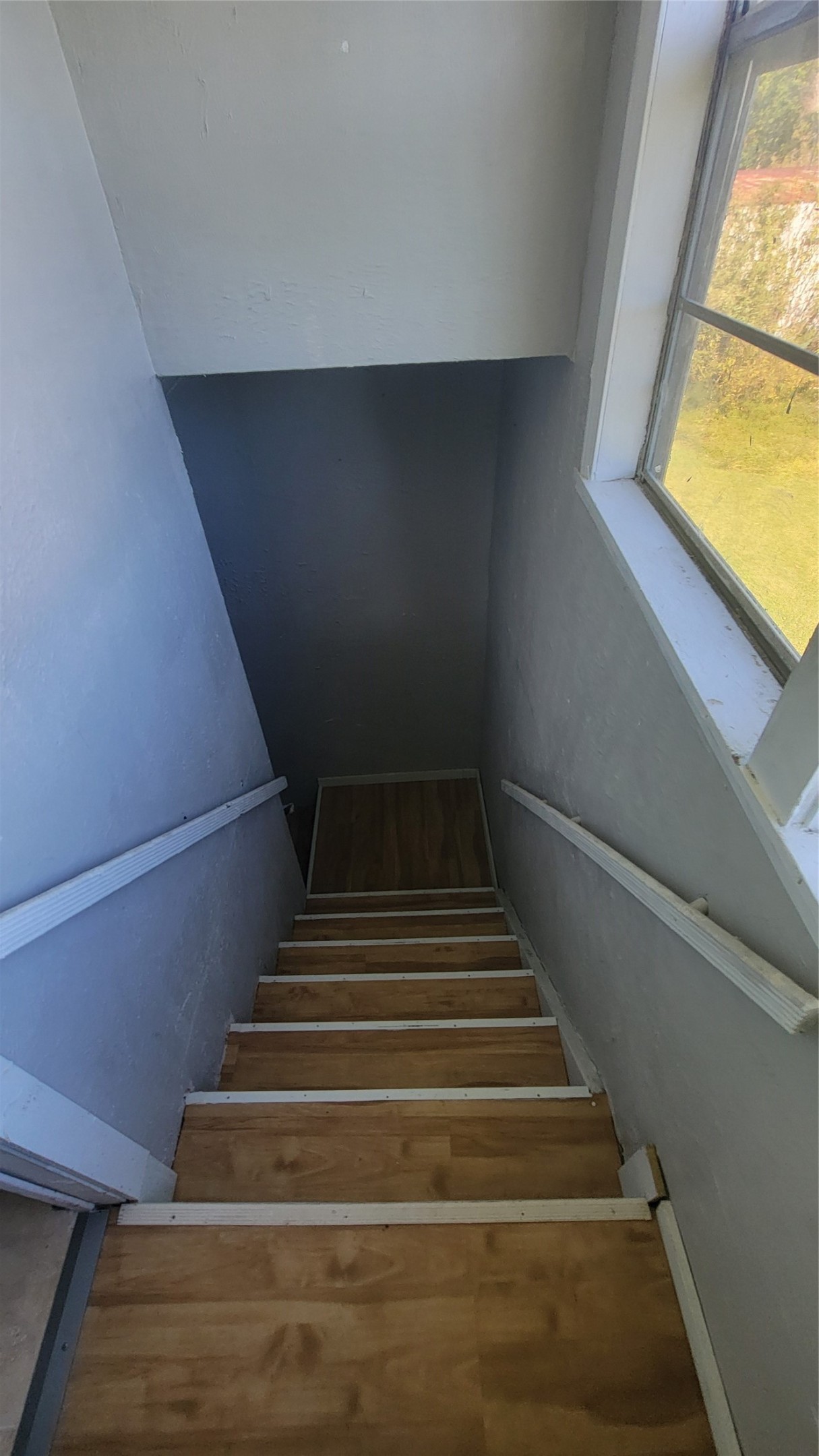 This photo shows a narrow staircase with wooden steps and white handrails, leading down to a lower level. There's a window on the right, providing natural light.