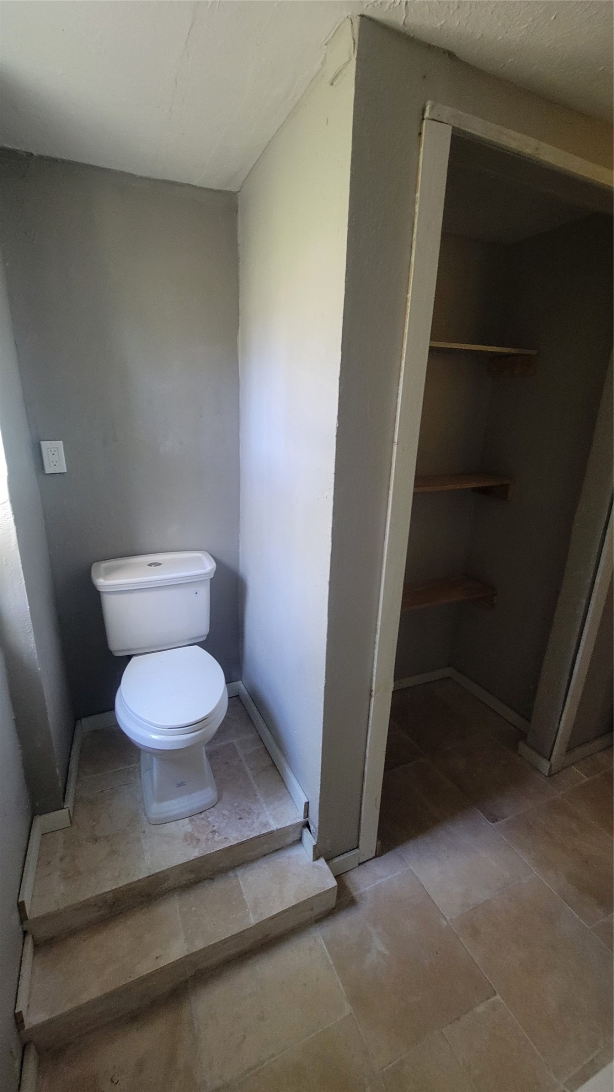 This photo shows a small bathroom area with a raised toilet and tiled flooring. There's an adjacent open closet space with built-in shelves. The walls are painted in a neutral color.