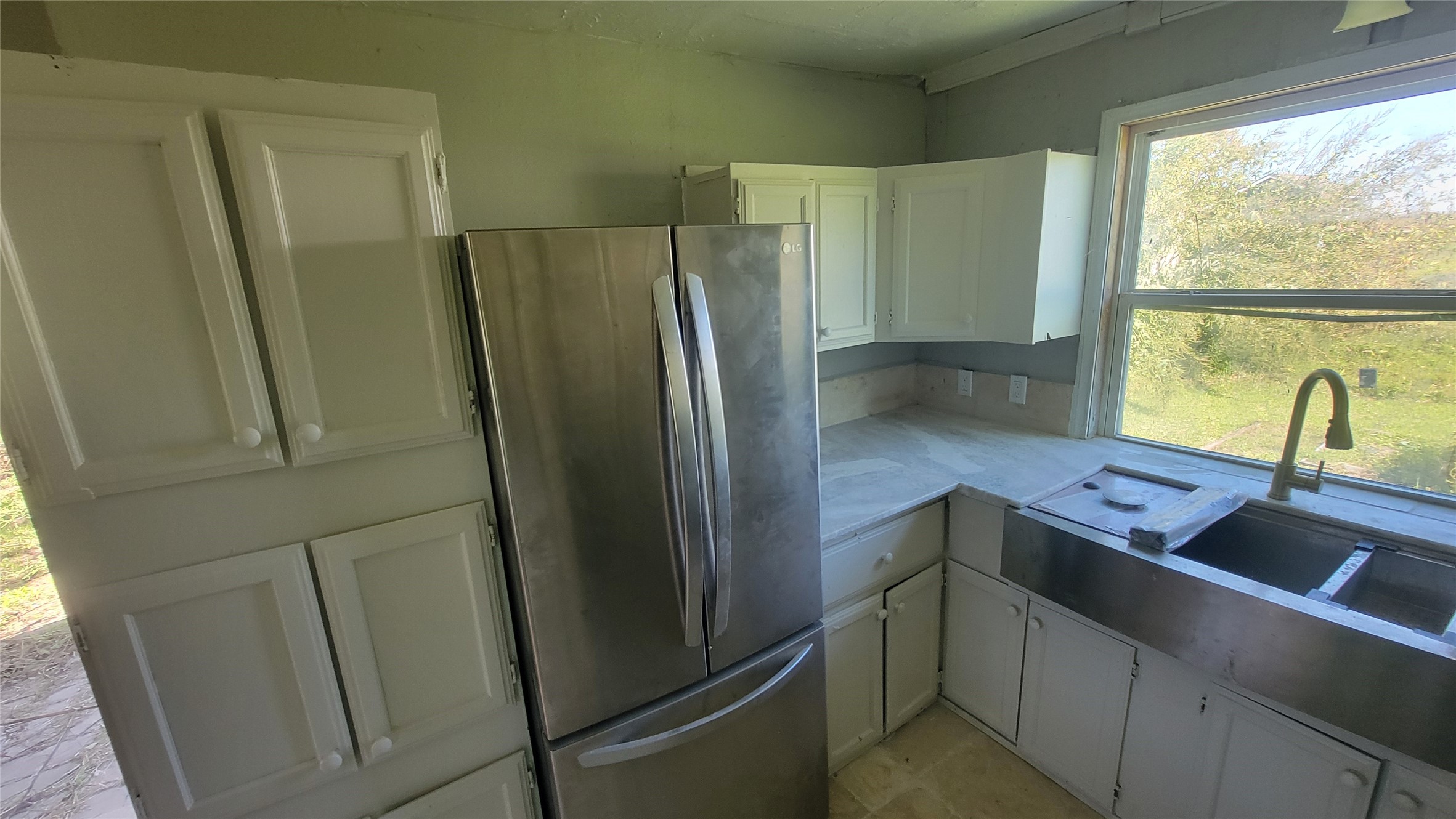 This kitchen features white cabinetry, a stainless steel refrigerator, and a large window providing natural light. The space includes a double sink with a view of the greenery outside.
