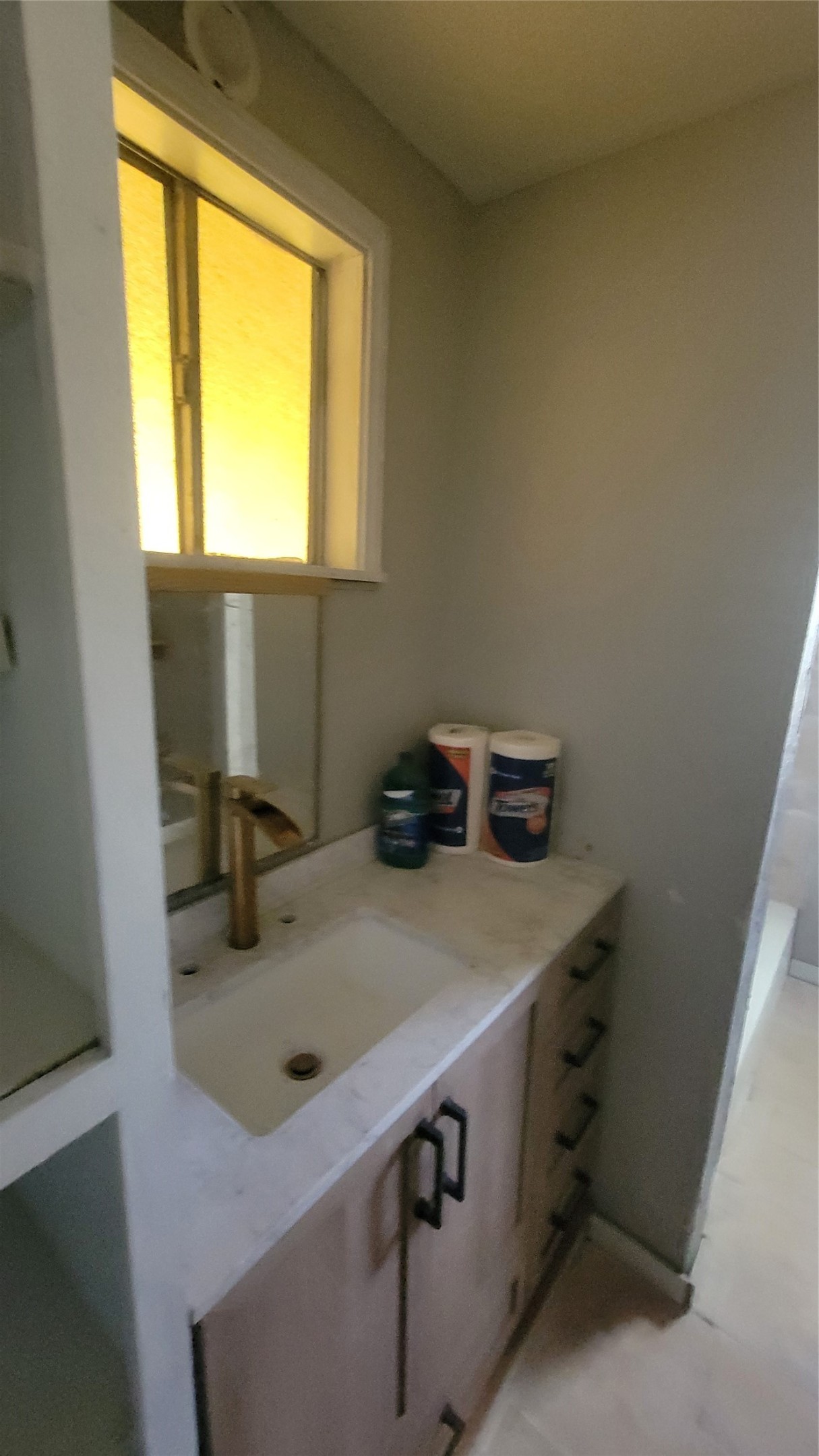 This photo shows a compact bathroom area featuring a sink with a marble countertop, modern brass faucet, and under-sink storage. A small window provides natural light, and there's shelving for additional storage.