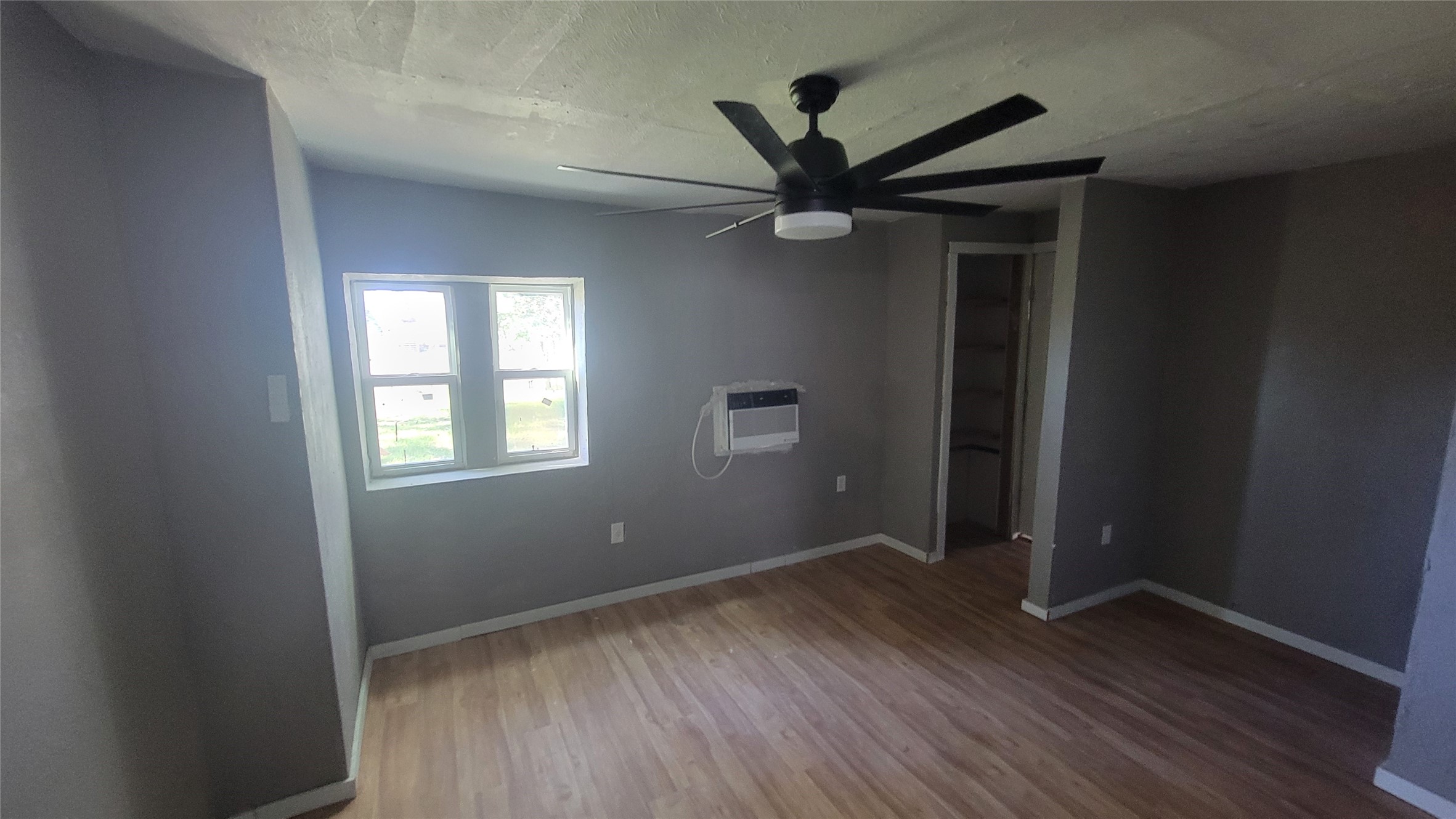 This room features neutral-toned walls, a large ceiling fan, and a window for natural light. It includes an AC unit on the wall and a small closet area, with light wood flooring throughout.