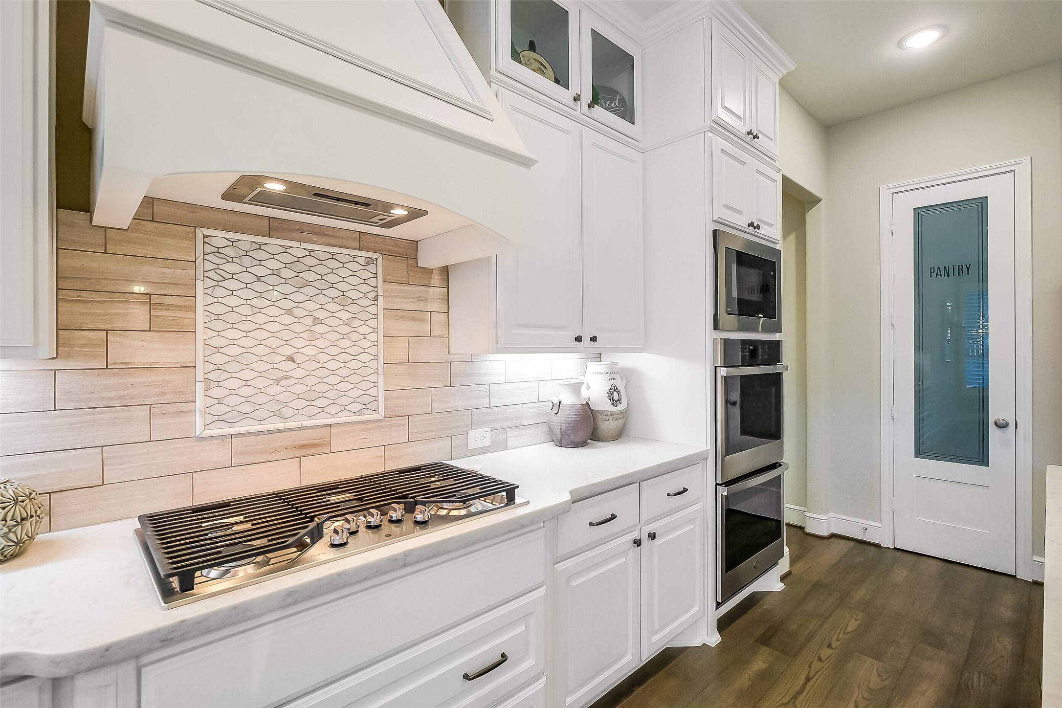 The heart of this kitchen's cooking wall features a gas cooktop, built-in double ovens, and a statement tile backsplash with a decorative inset. A large walk-in pantry just beyond keeps everything organized and within reach.