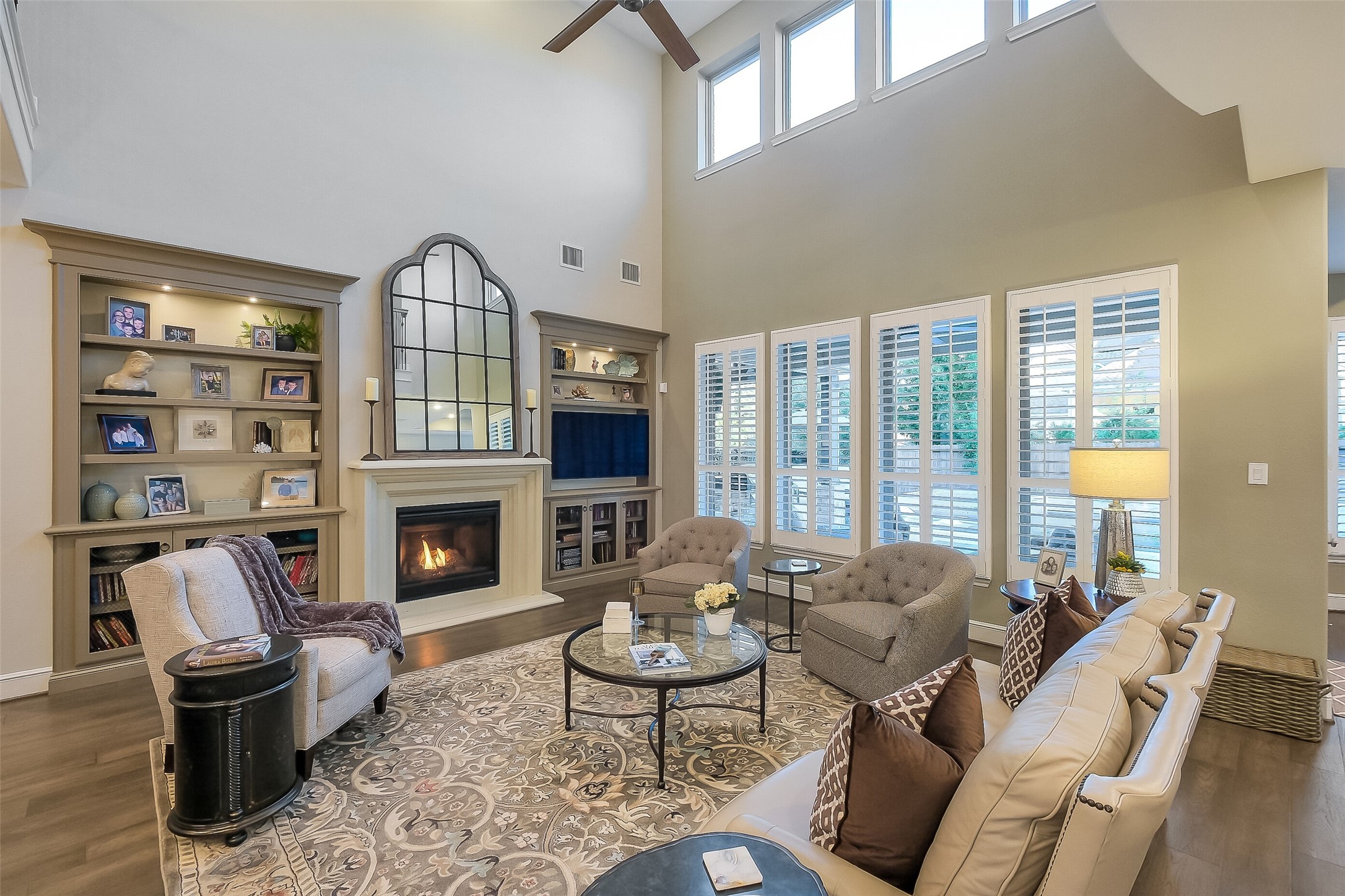 Soaring two-story ceilings and a wall of plantation-shuttered windows fill this living room with natural light. A cozy gas fireplace and custom built-in shelving create a warm, welcoming gathering space. Soaring two-story ceilings and a wall of plantation-shuttered windows fill this living room with natural light. A cozy gas fireplace and custom built-in shelving create a warm, welcoming gathering space.