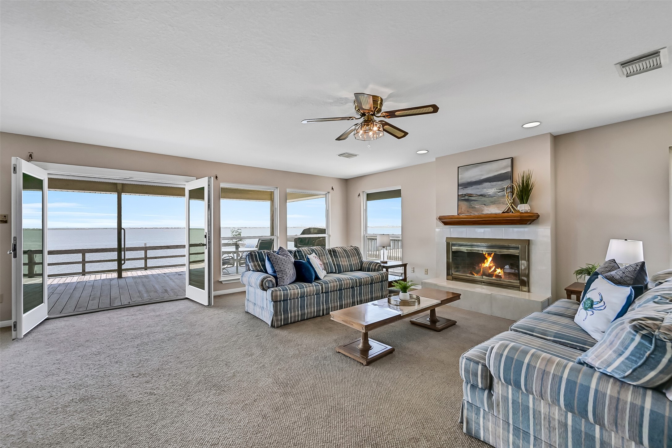 The living room is truly the heart of the home, featuring a wood-burning fireplace with gas starter, fresh neutral paint, and walls of windows that frame breathtaking panoramic views of West Bay. French doors open directly onto the expansive waterfront deck.