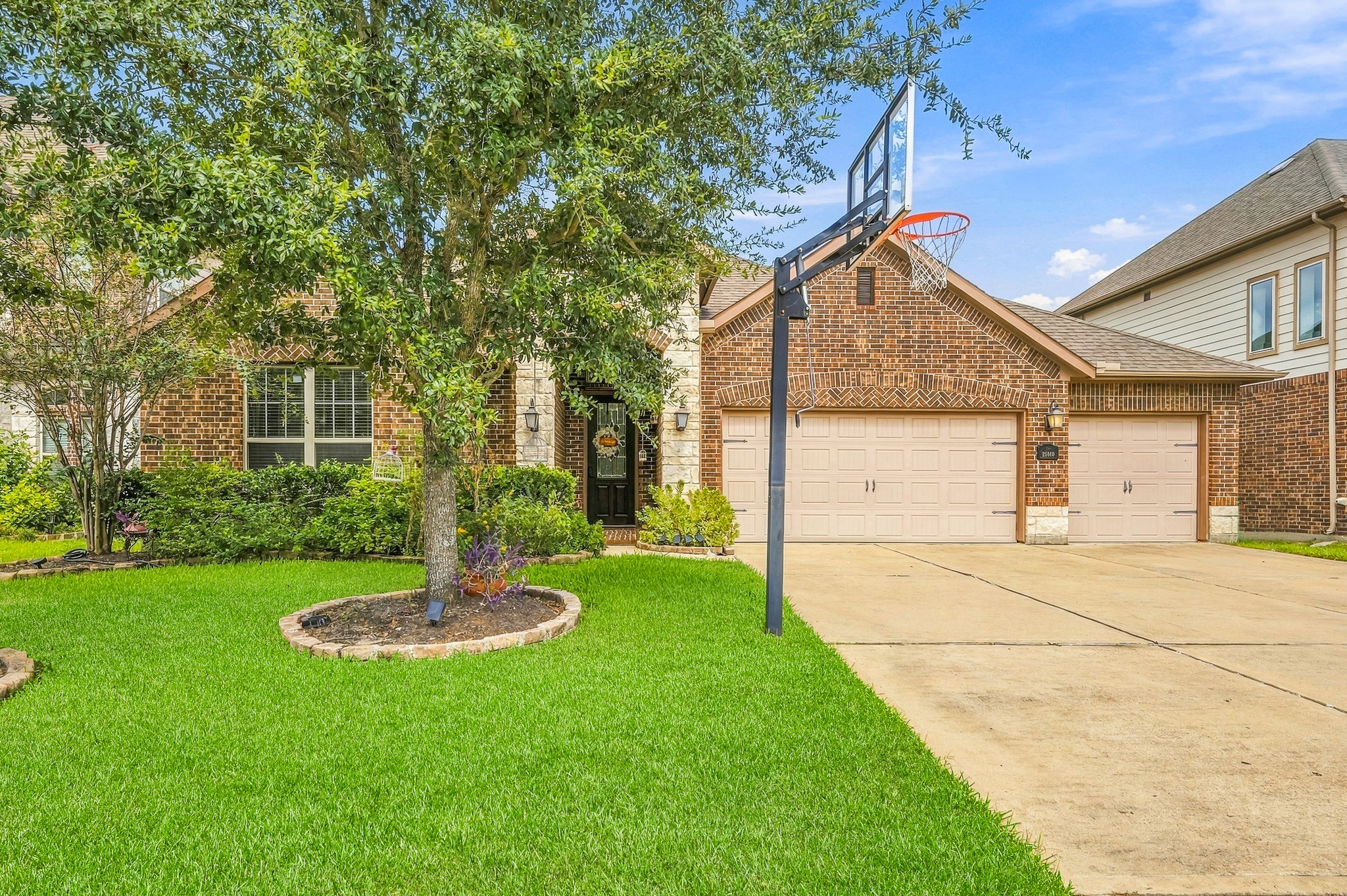 Charming brick home featuring a spacious three-car garage, lush landscaping, and a basketball hoop for outdoor fun.