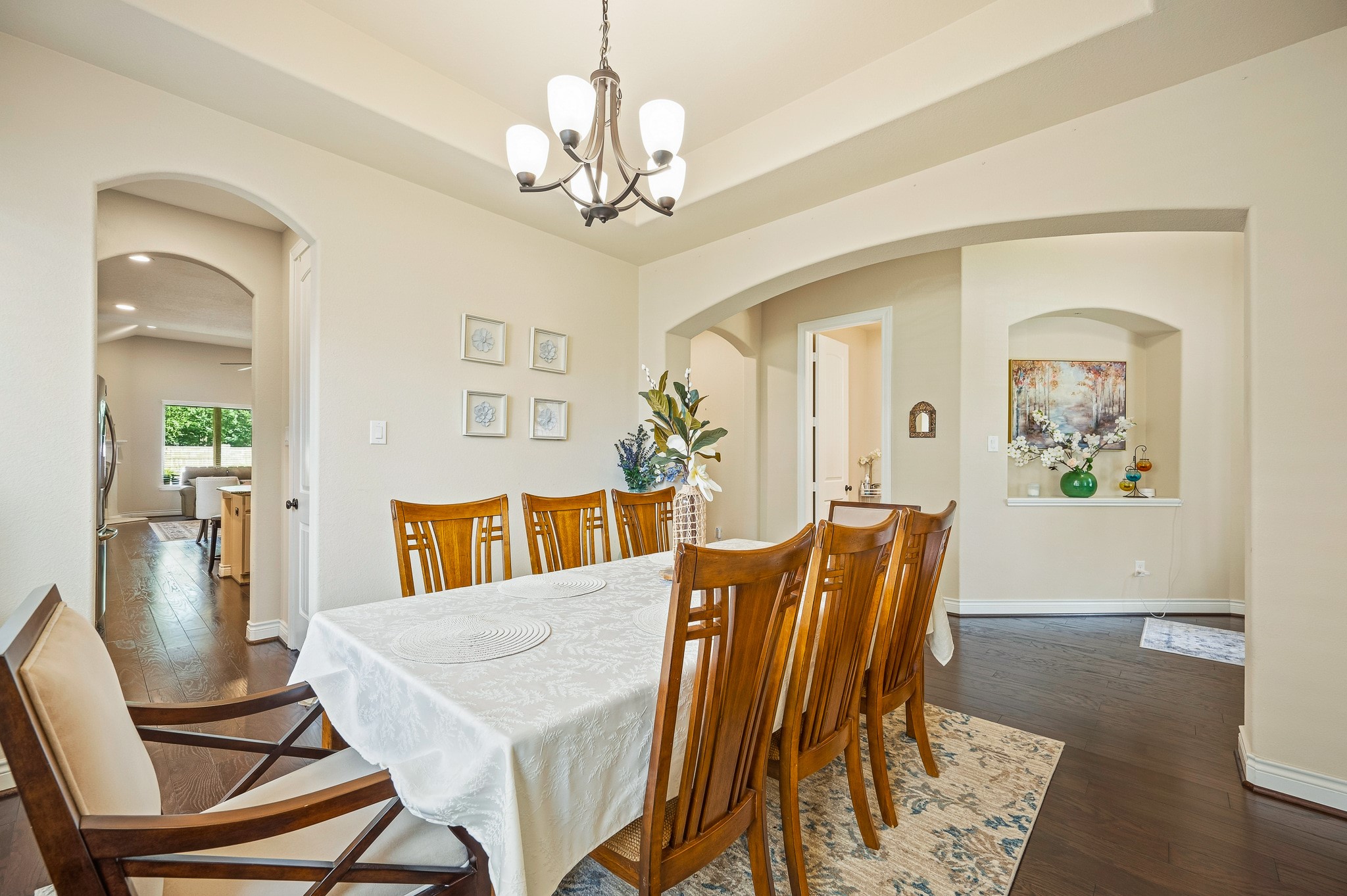 Experience the elegance of a dining room featuring a long wooden table with a white tablecloth and matching chairs beneath a stylish chandelier, seamlessly connected to open kitchen and living spaces through graceful archways, enhancing the home's flow and charm.