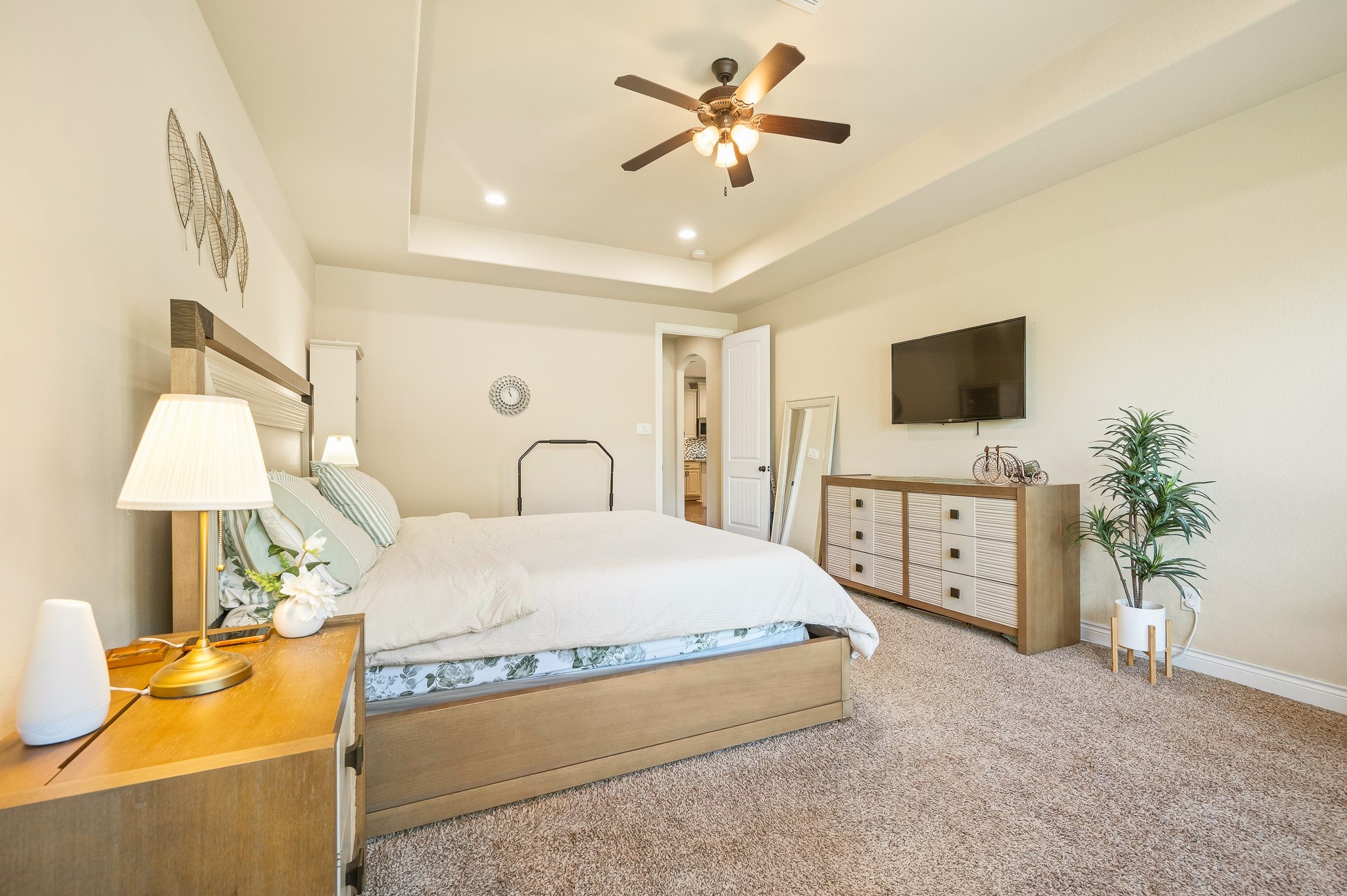 A spacious bedroom with plush carpeting and abundant natural light, featuring a comfortable bed, elegant ceiling fan, modern dresser, and mounted TV for an inviting retreat.