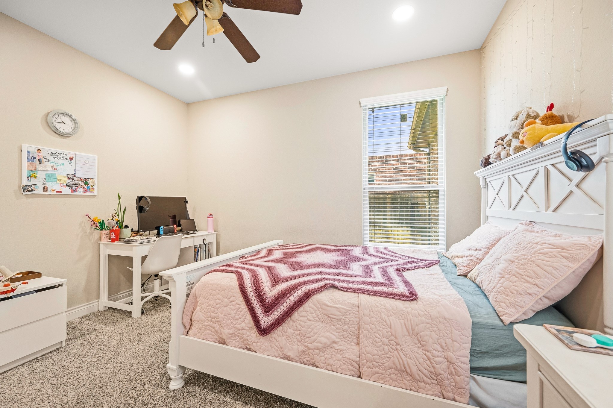 Charming bedroom with soft neutral tones, a cozy bed, and a functional workspace under natural light from a large window.