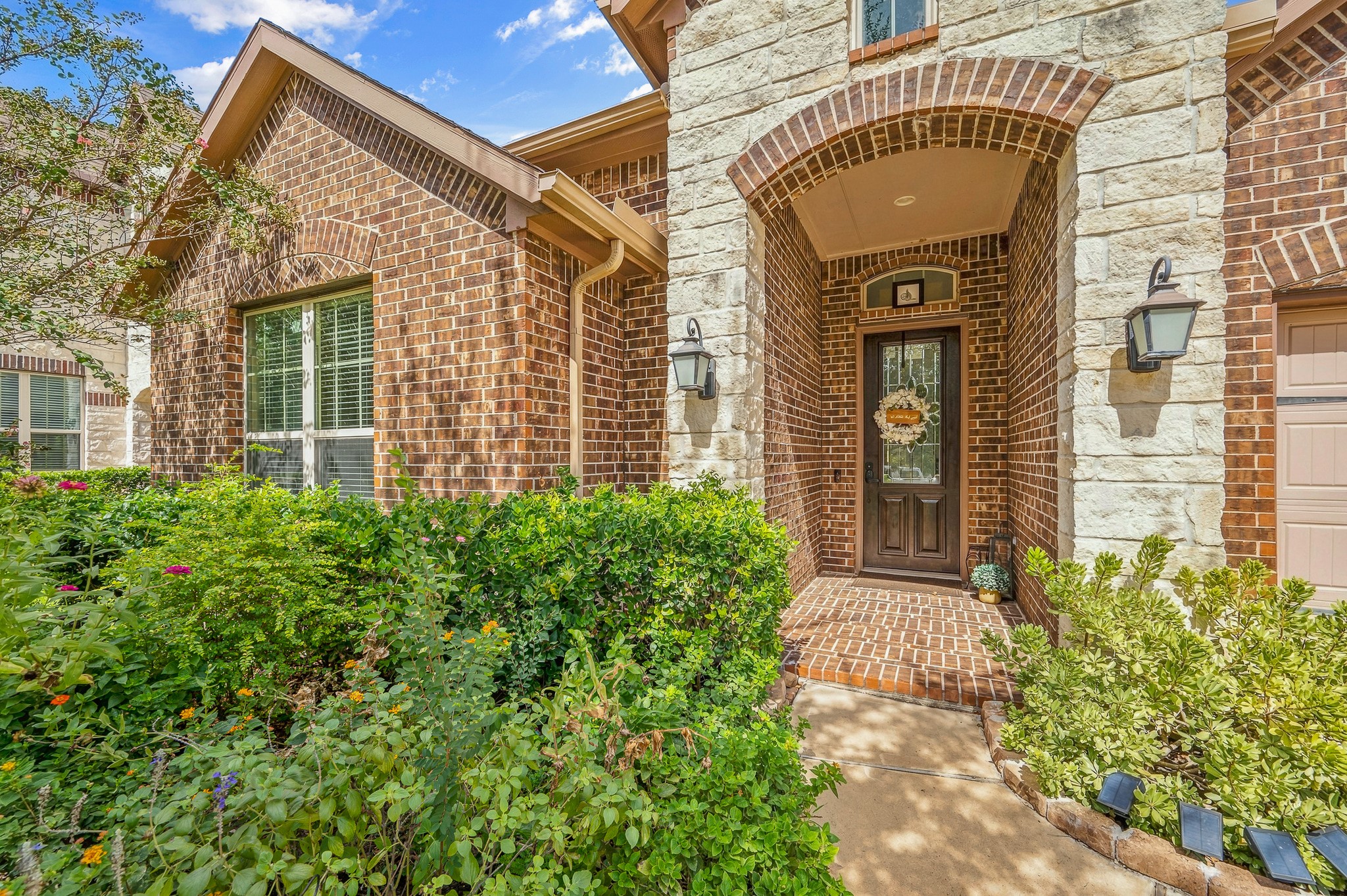 Charming brick exterior with a welcoming arched entryway, surrounded by lush landscaping and enhanced by decorative lanterns for a warm, inviting atmosphere.