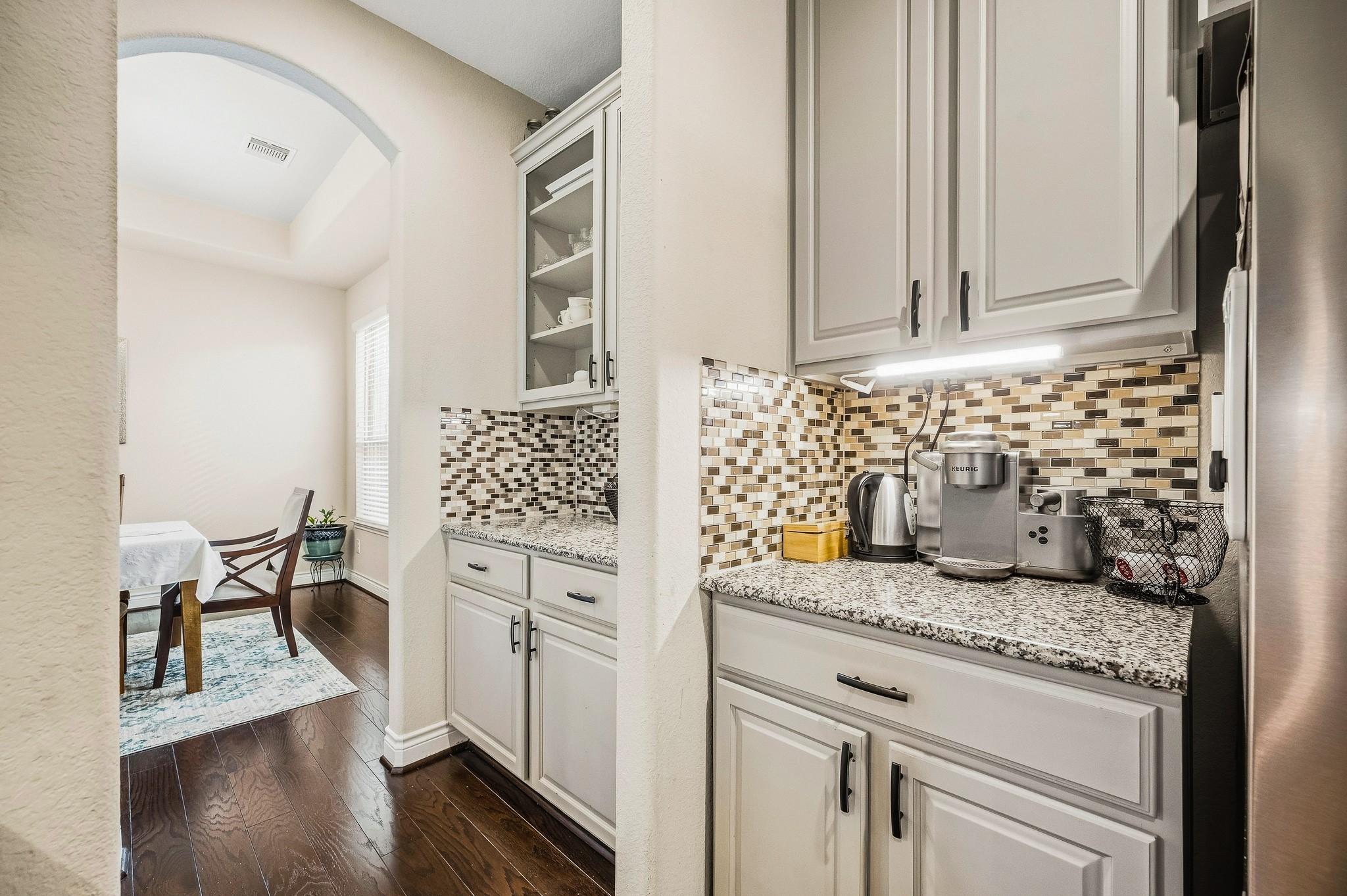 Experience the charm of an elegant kitchen nook with sleek granite countertops and a stylish mosaic tile backsplash, seamlessly flowing into a bright dining area adorned with rich hardwood flooring.