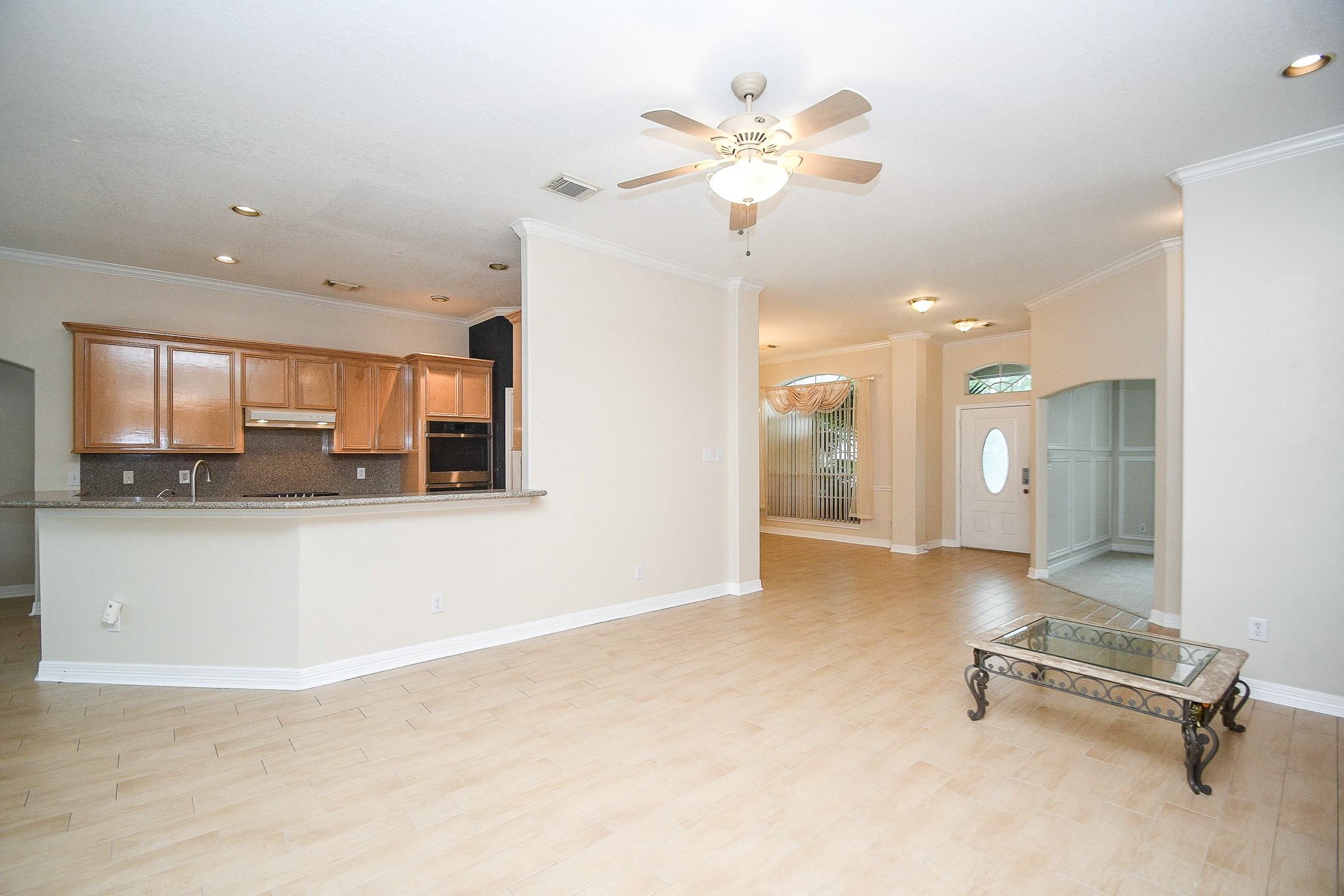 This open-concept living space features a bright kitchen with wood cabinets and a breakfast bar, flowing into a spacious living area with tile flooring, a ceiling fan, and abundant natural light. The front entry is welcoming, with a decorative glass door and an adjacent room. Perfect for entertaining and family gatherings.